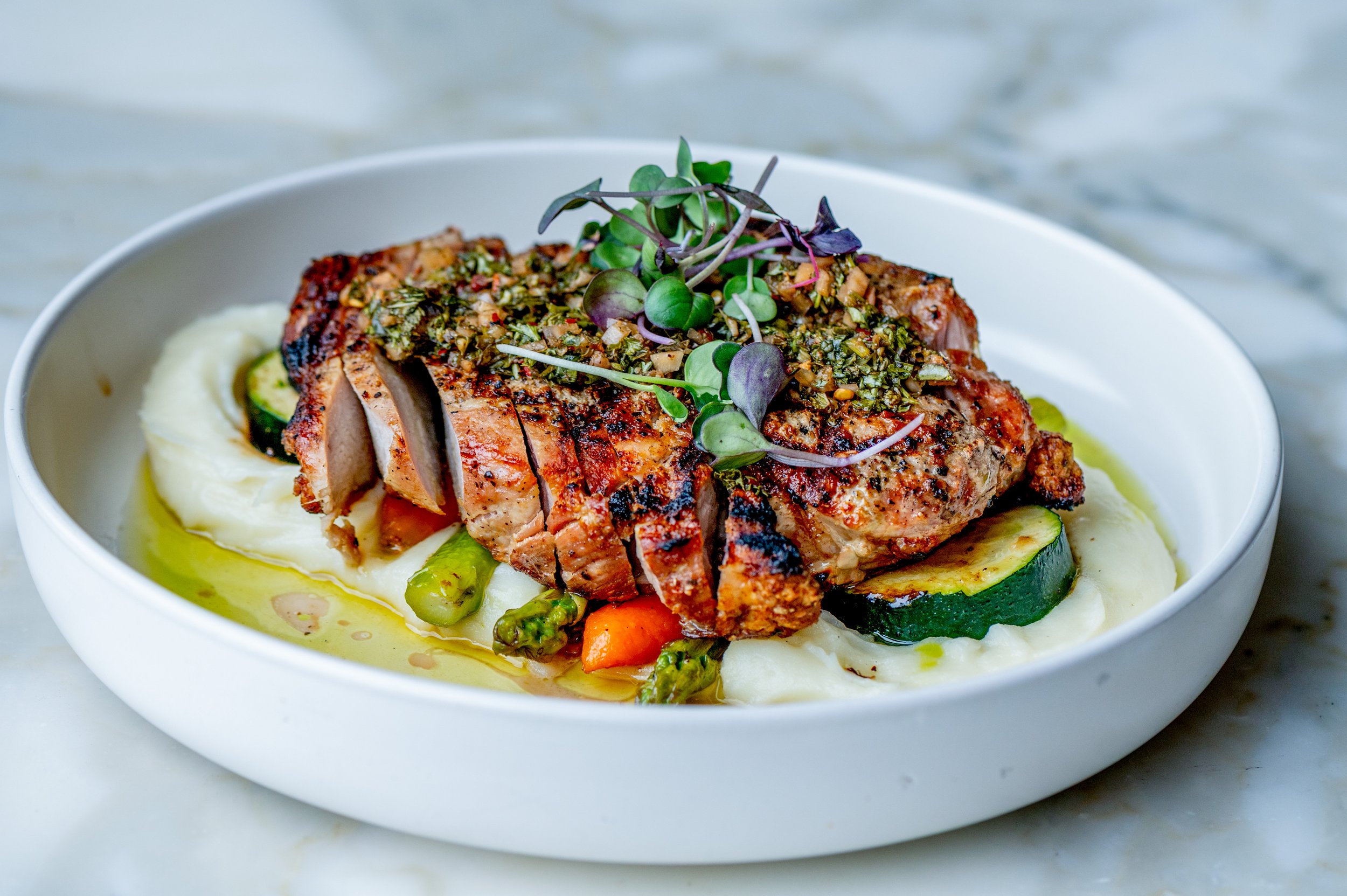 A plated meal with grilled salmon topped with herbs and microgreens, served over mashed potatoes with green beans and carrots, garnished with a drizzle of olive oil.