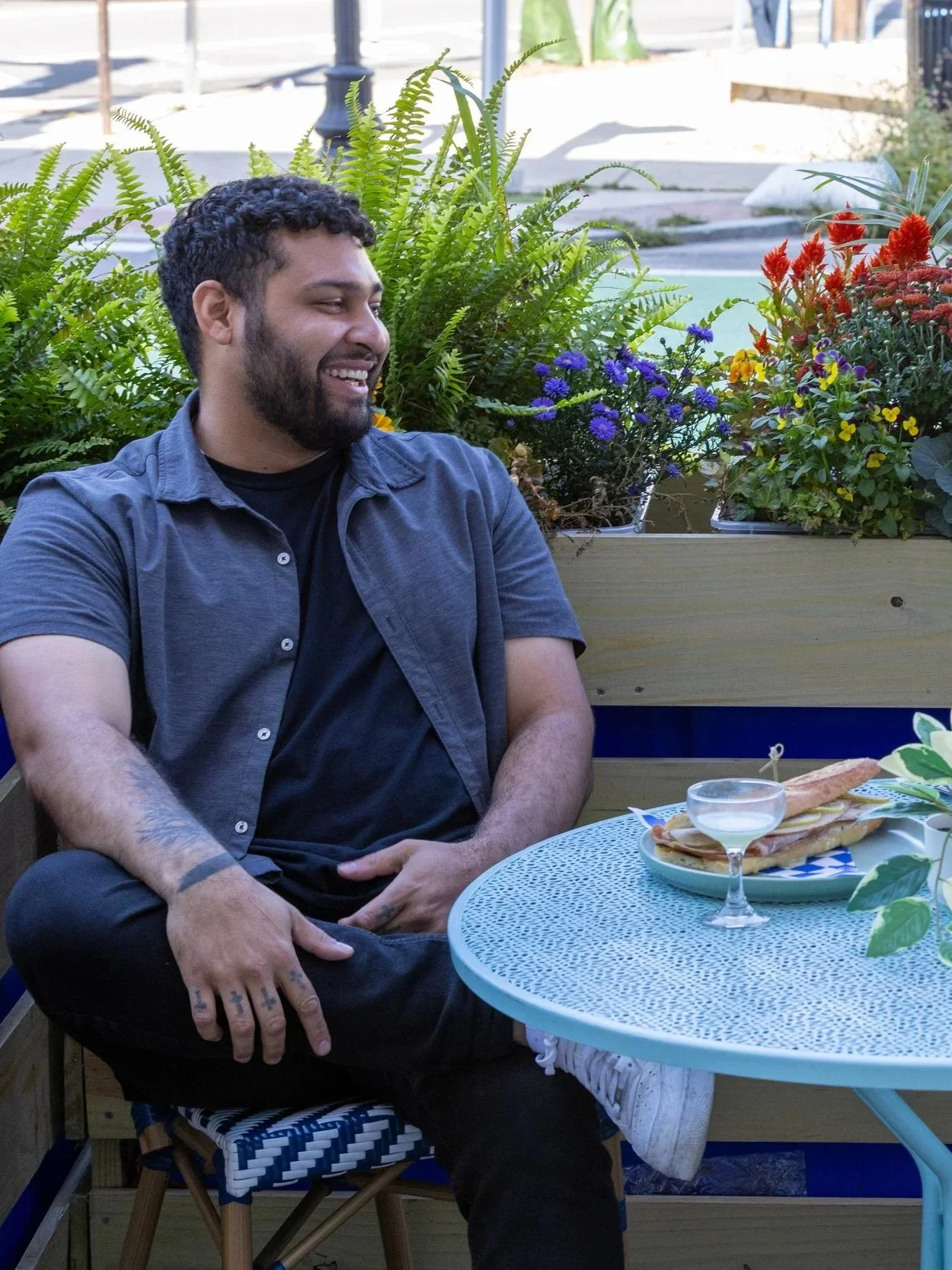 A man with dark curly hair and a beard sitting outdoors at a round table with a plate of bread and a cocktail glass, smiling and looking to the side, surrounded by green plants and colorful flowers.