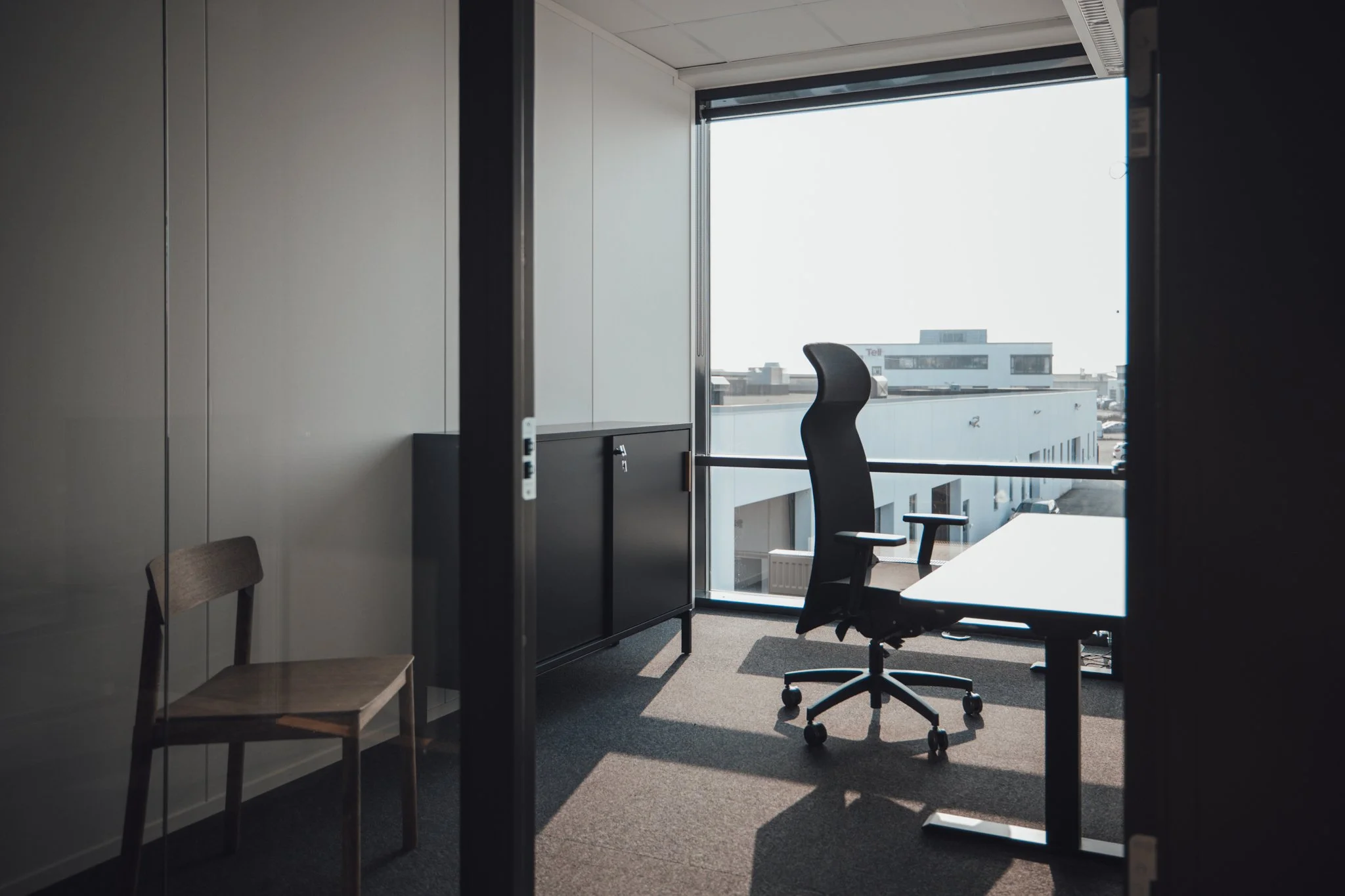 Kontor med skrivebord, kontorstol, og et stort vindu som viser bygninger utenfor.