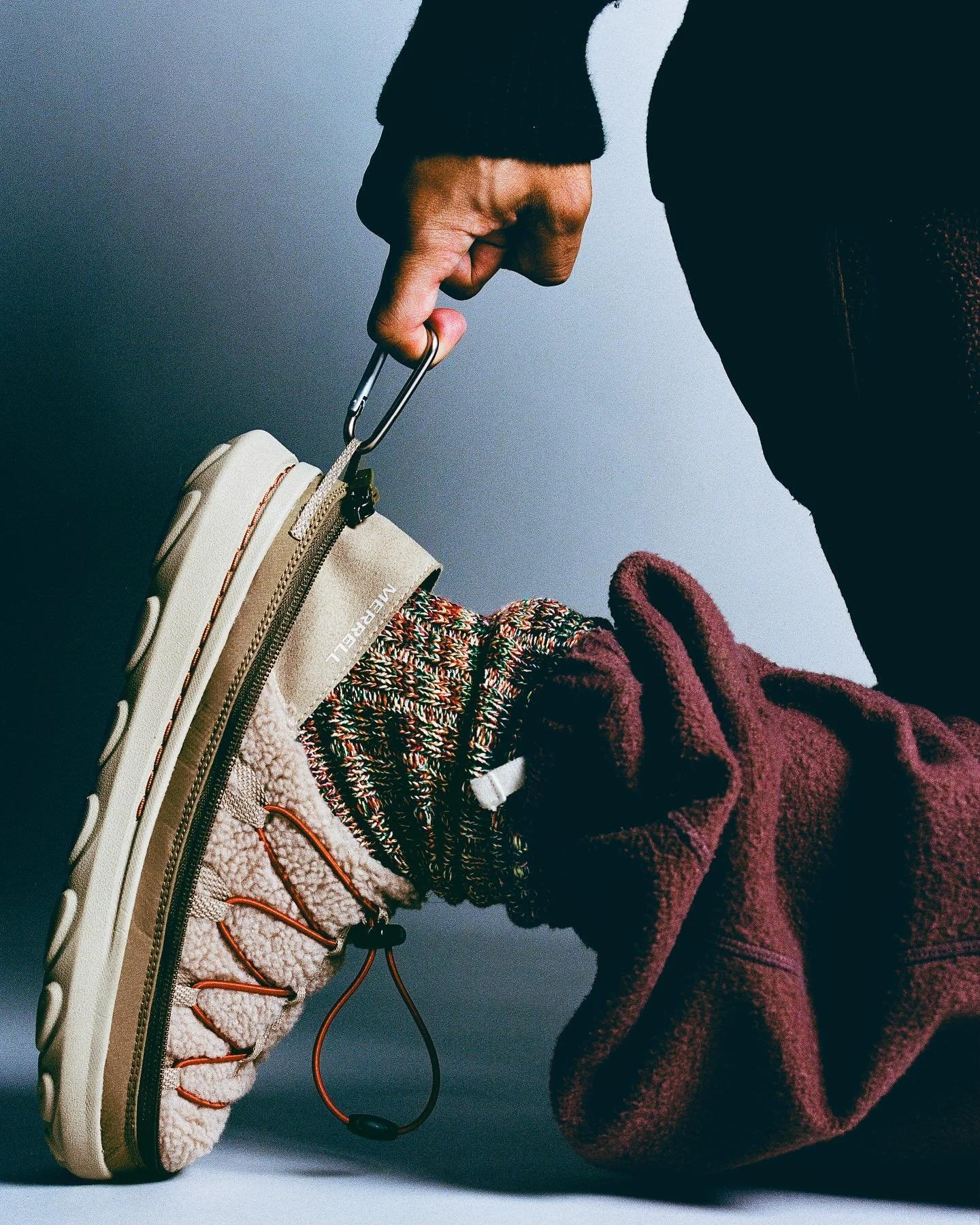 A close-up of a person’s foot wearing a beige sneaker with a shearling lining and orange accents, being held by a hand with a carabiner, against a plain background.