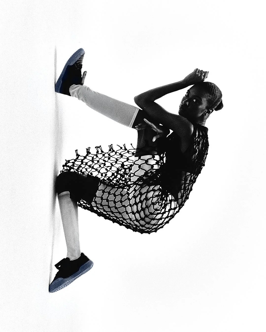 A young girl wearing a black netted dress, white leggings, and black sneakers is sitting on the floor, holding a pose with one hand behind her head and the other across her chest, against a plain white background.