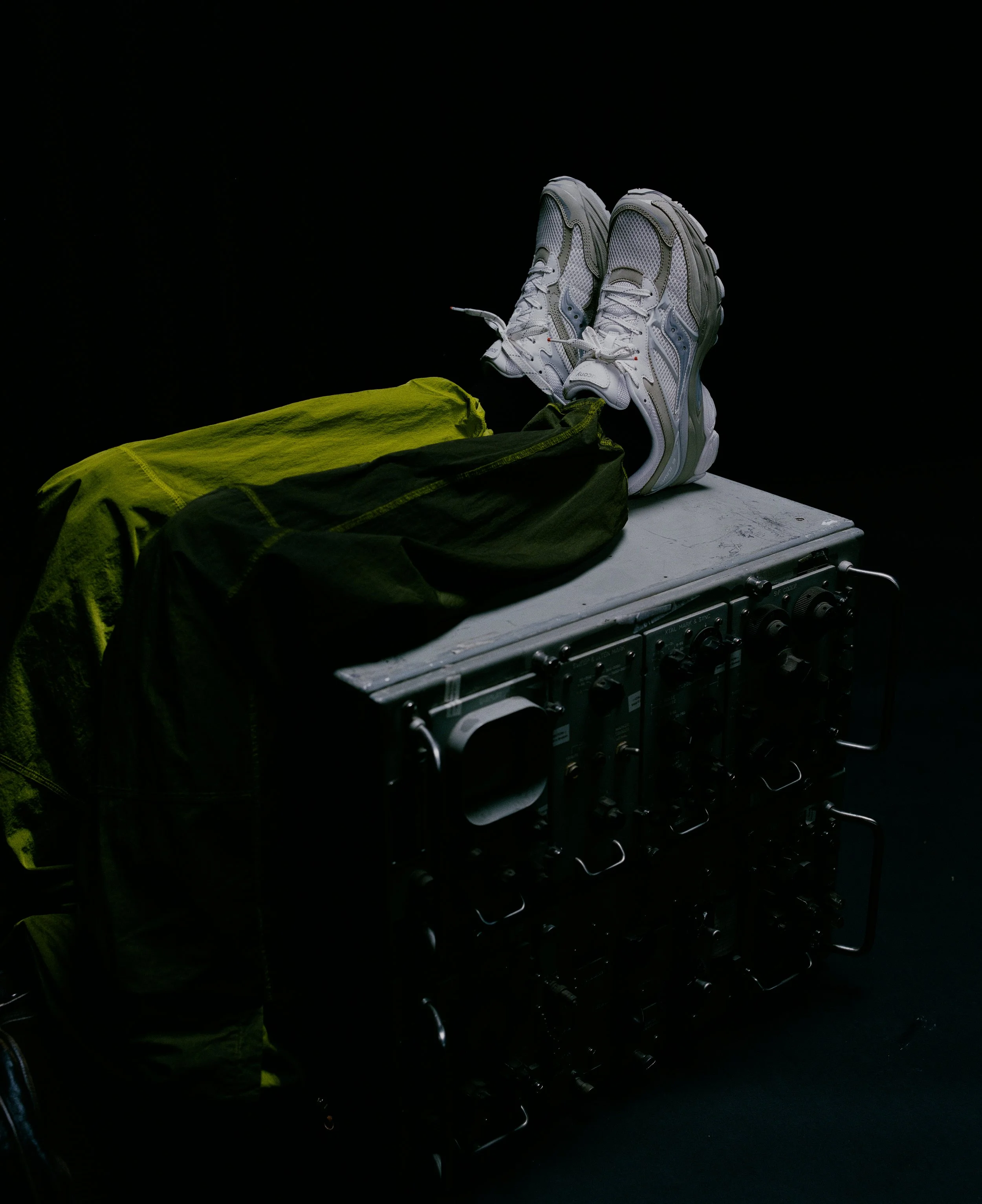 A pair of white athletic shoes, a yellow jacket, a black jacket, and a metallic suitcase against a black background.
