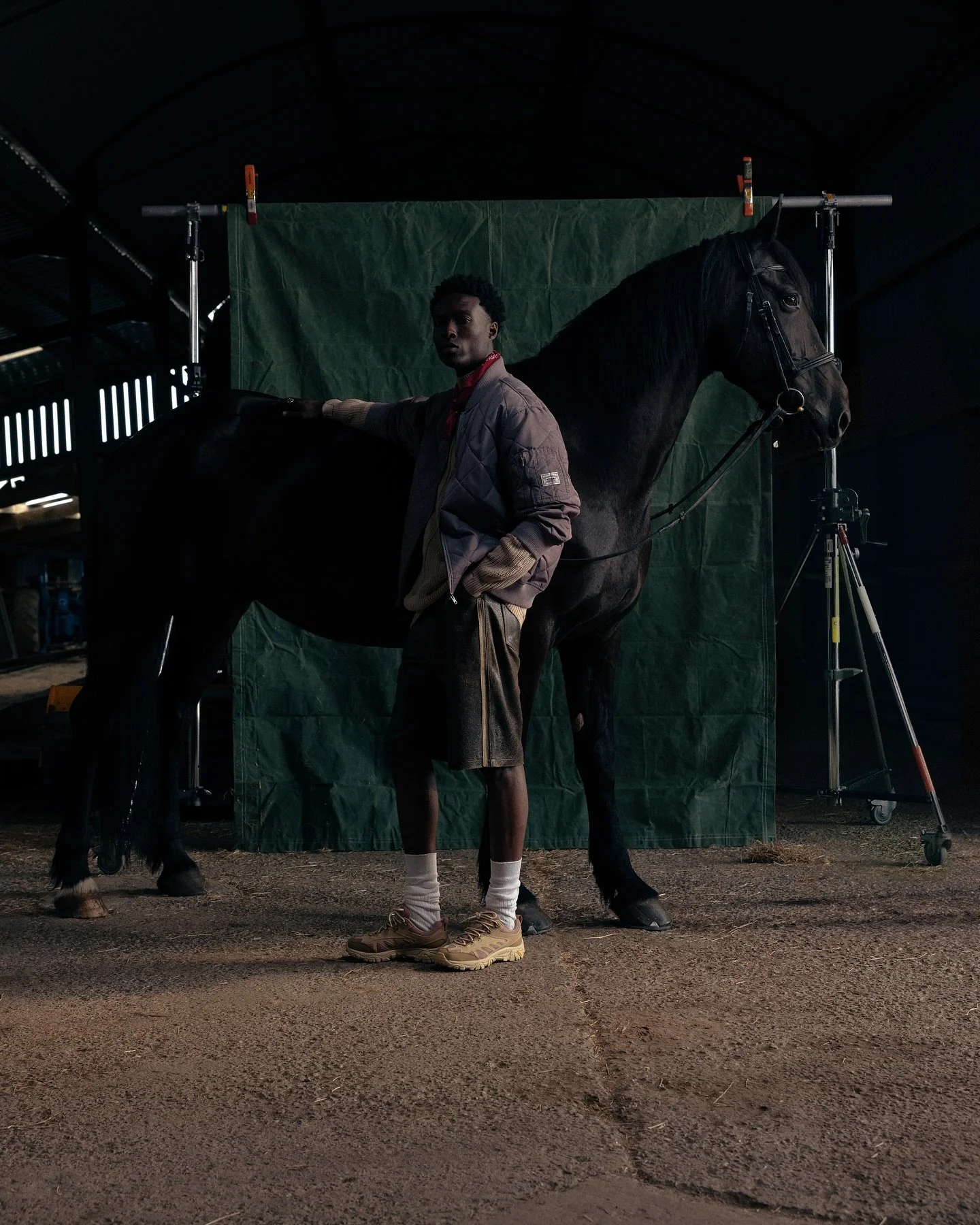 A young man standing next to a black horse inside a dark stable or indoor area, with a green backdrop behind the horse.
