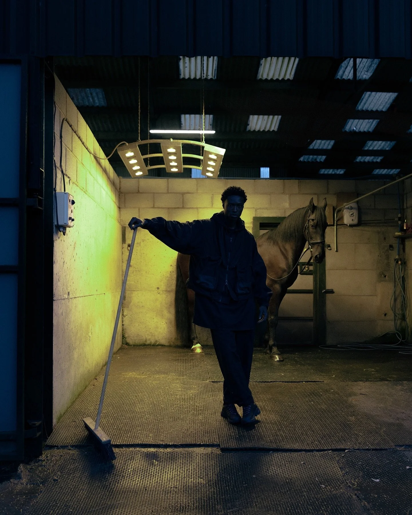 A person in dark clothing standing in a stable or grooming area with a horse in the background, holding a broom leaning against the wall, under fluorescent lights.