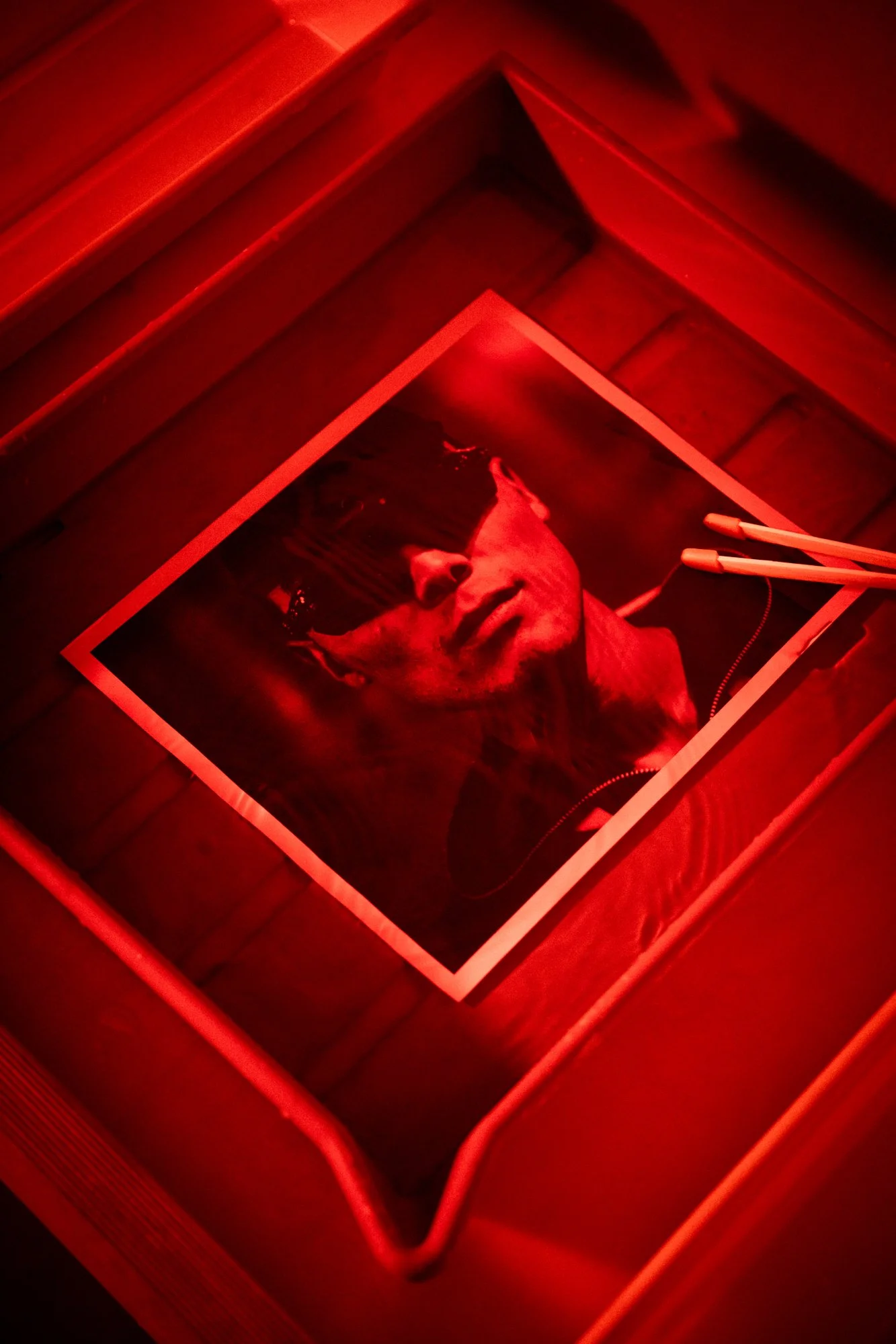 A glossy photograph of a man with dark hair, wearing sunglasses and headphones, illuminated by red lighting, lying on a wooden surface.