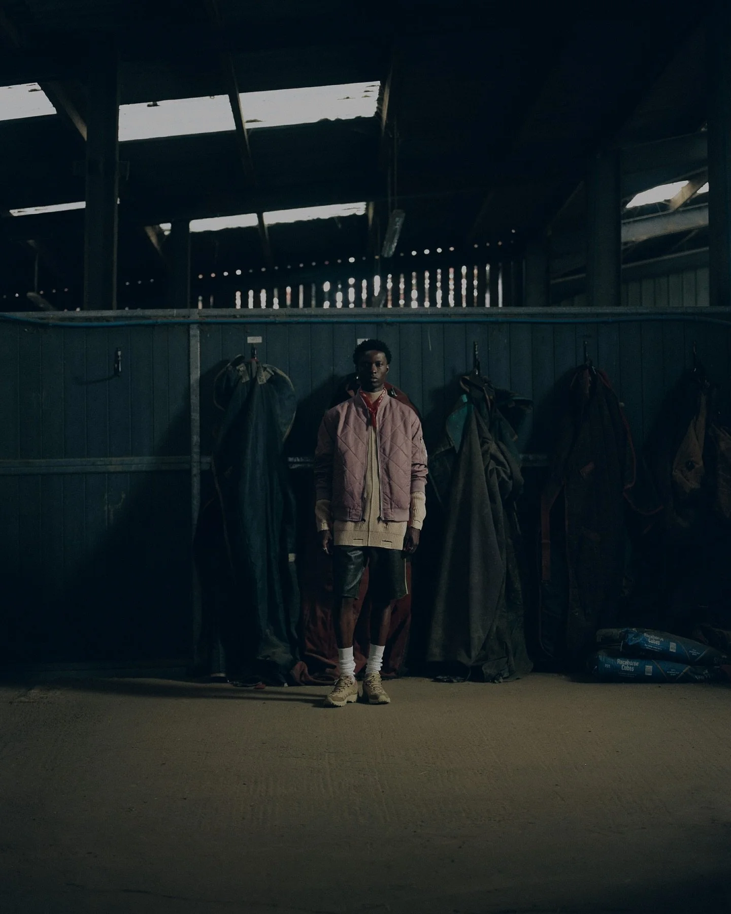 A young man standing in a dimly lit stable or horse barn, surrounded by hanging riding gear such as saddles and bags, with a wooden wall and ceiling in the background.