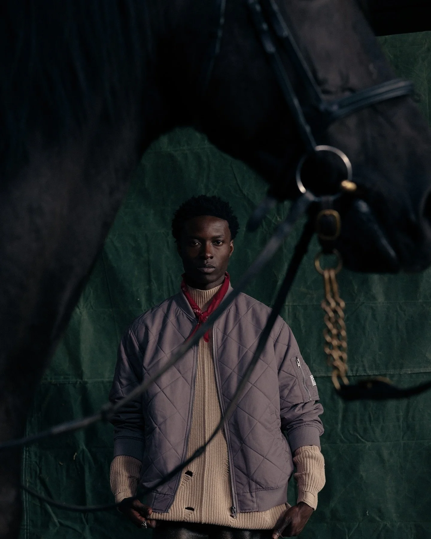 A young man with dark skin and curly hair standing against a green backdrop, wearing a tan sweater and a brown quilted jacket, appears to be in a barn.