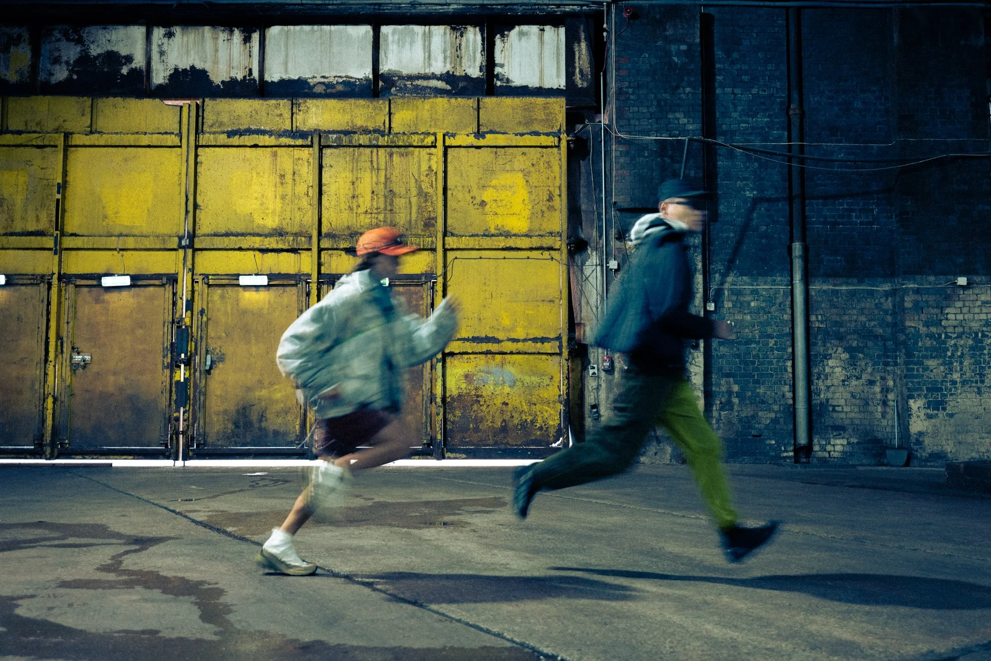 Two people jogging outdoors in front of a large, yellow industrial-style gate in an urban setting.