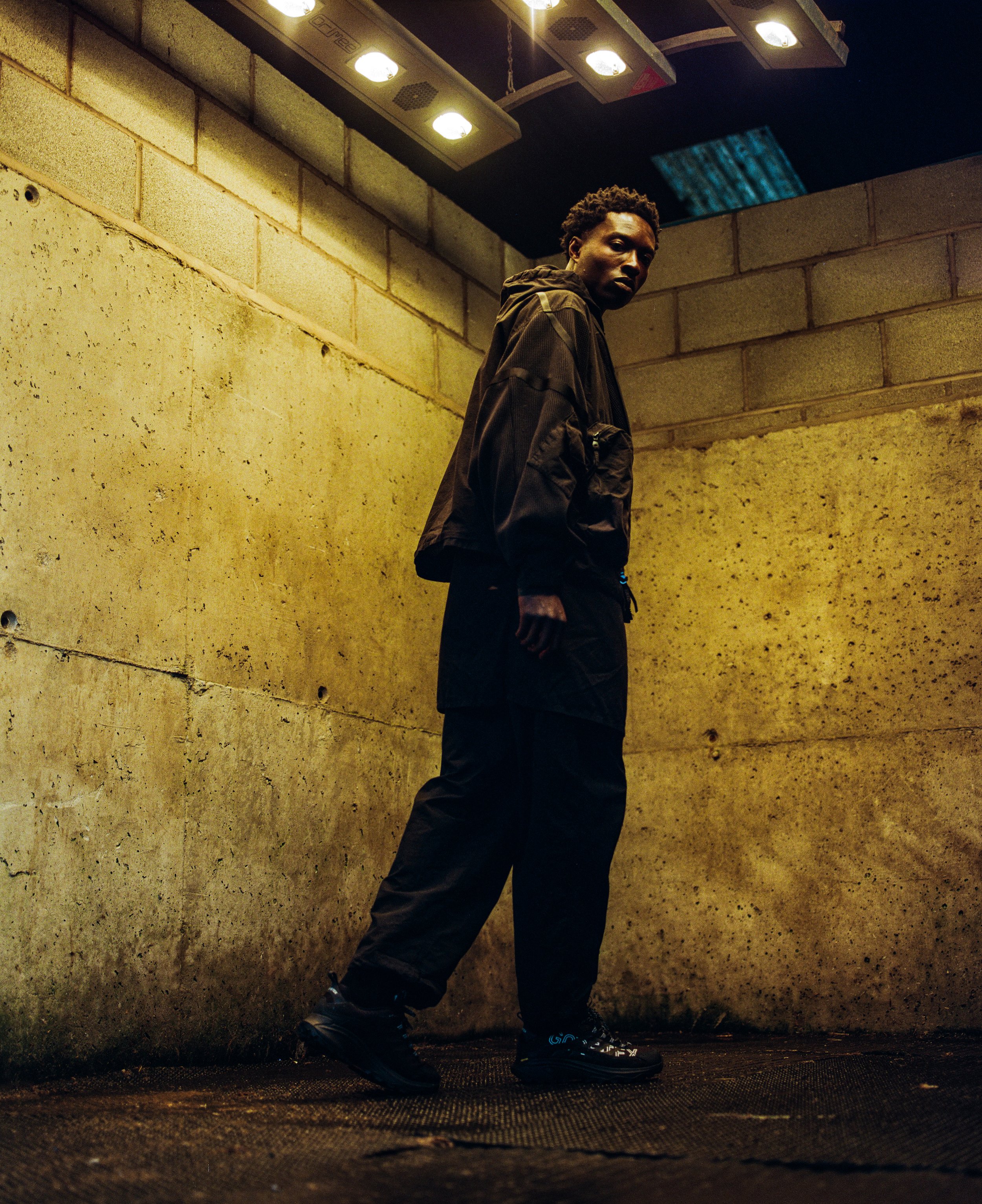 A man standing in a dimly lit, industrial-style space with concrete walls, wearing dark clothing and black shoes, looking at the camera.