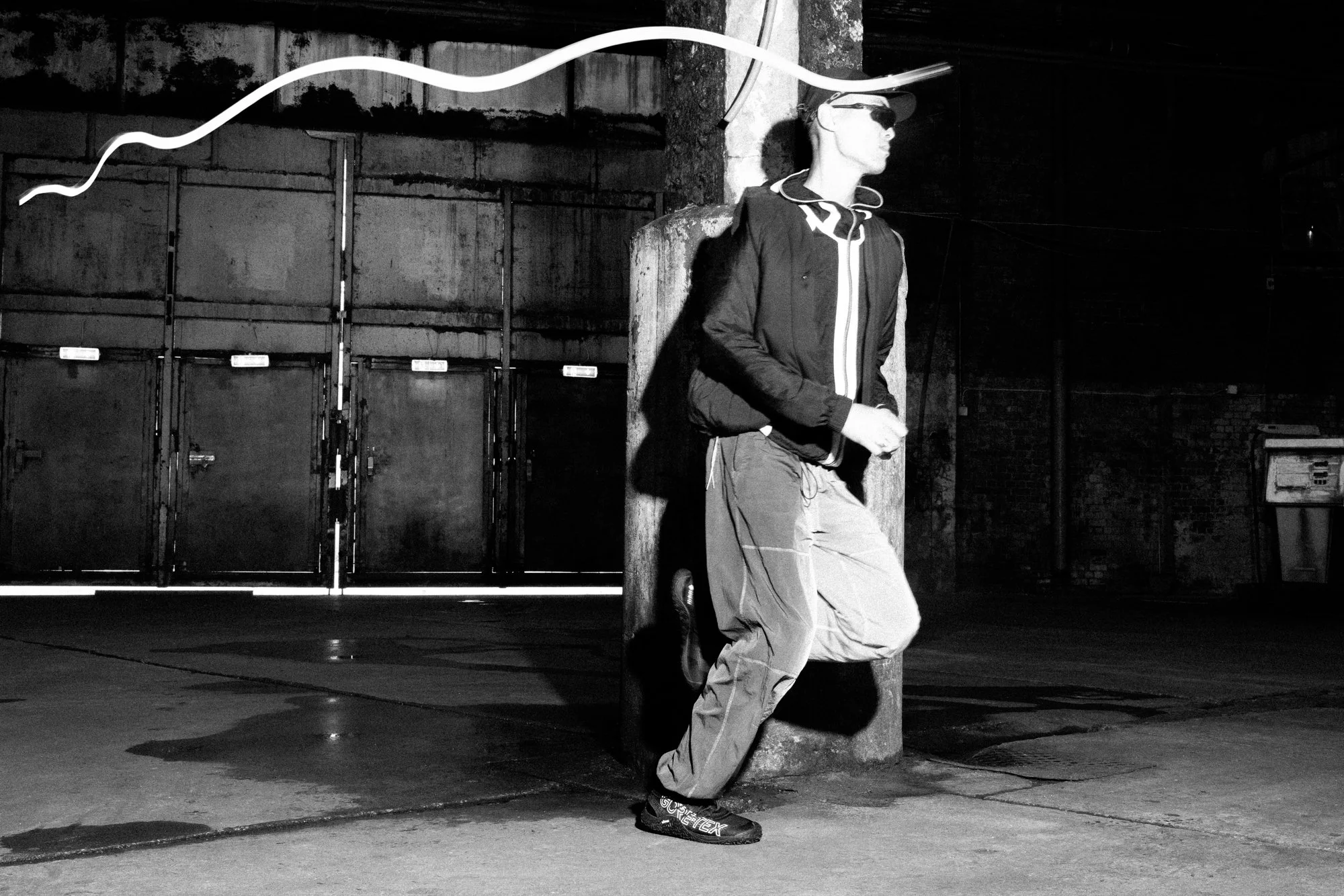 A person wearing a cap and glasses, leaning against a utility pole, walking in an urban alley at night, with a brick wall and metal storage units in the background, in black and white.