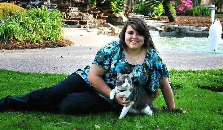 A woman lying on grass in a park with a gray and white cat, smiling at the camera.