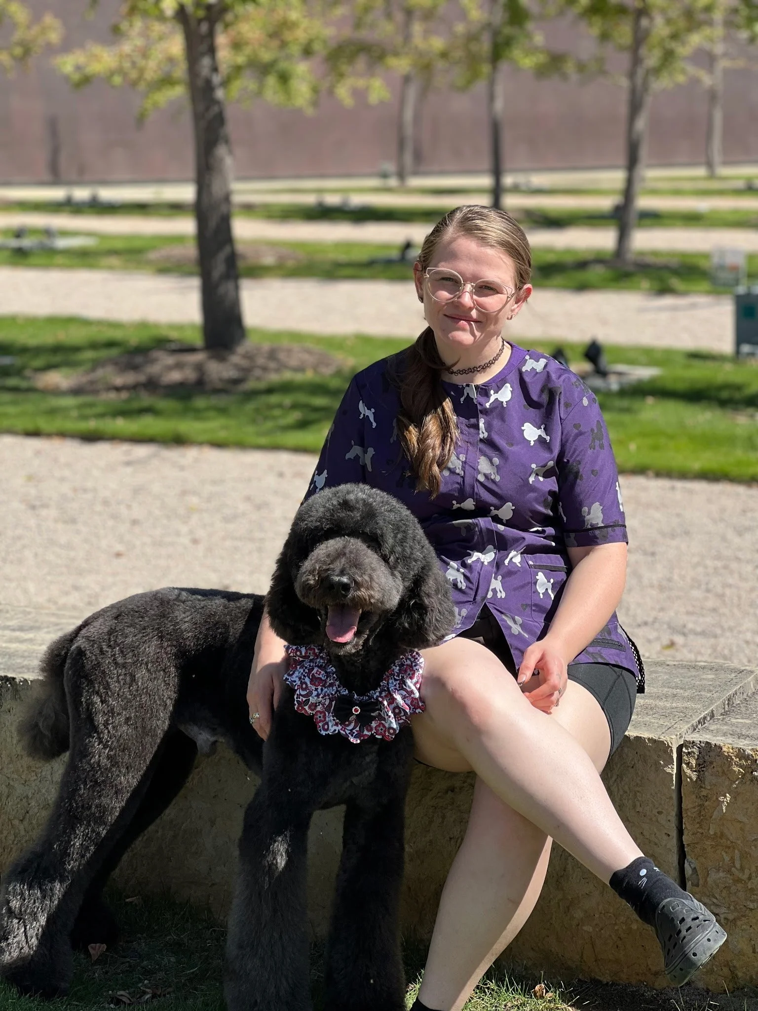 A girl with glasses and a purple shirt with dog illustrations, sitting on a stone bench in a park, smiling with a large black poodle sitting beside her, wearing a floral bandana. Trees and a pathway are visible in the background.