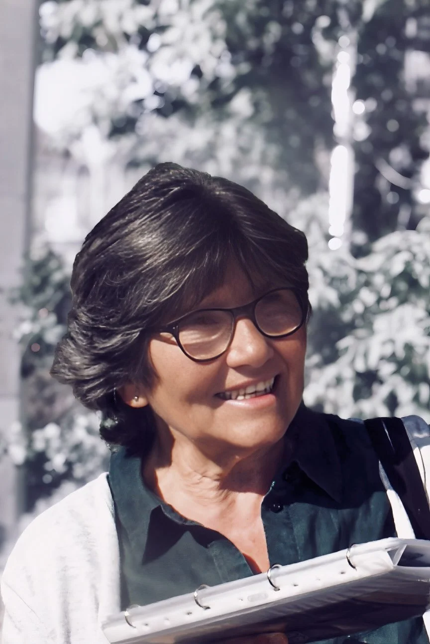 Eine lachende Frau mit kurzen dunklen Haaren und Brille hält ein aufgeschlagenes Notizbuch. Im Hintergrund ist eine Winterlandschaft mit verschneiten Bäumen zu sehen.
