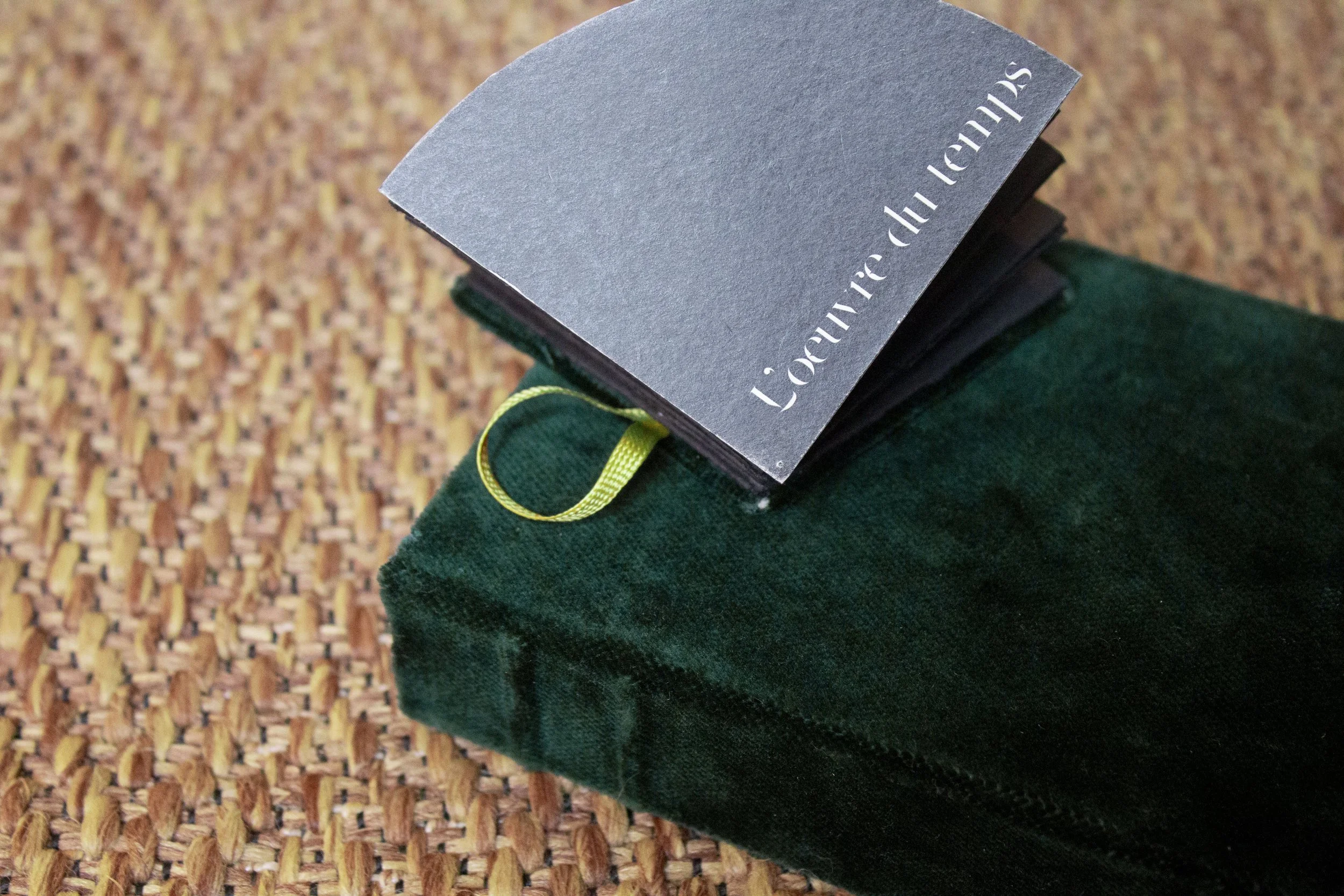 Un livre intitulé 'L'œuvre de l'écho' posé sur un étui en velours vert foncé, avec une étiquette verte attachée, posé sur un tapis tissé en fibres naturelles.