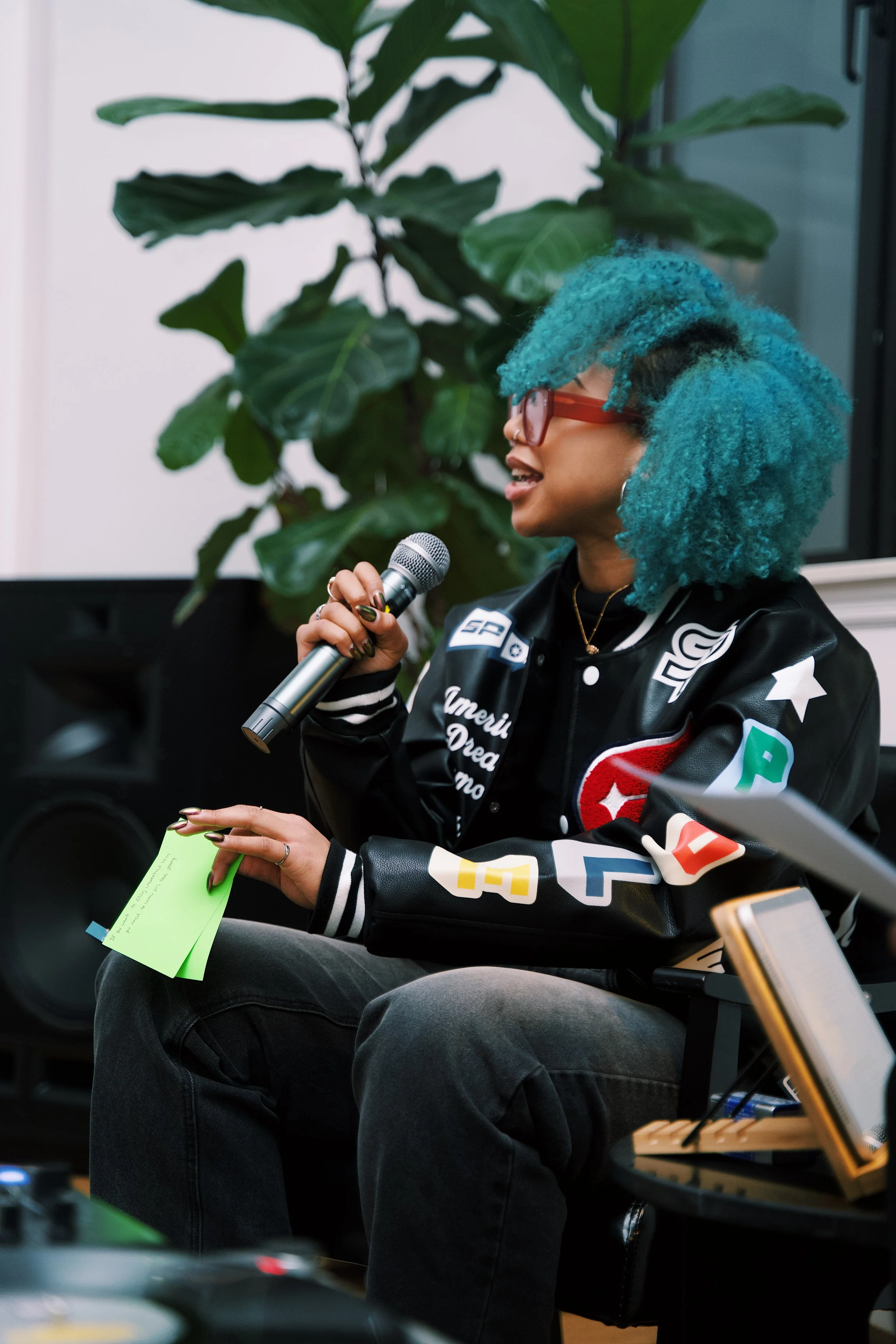 A woman with curly orange hair, glasses, and a nose piercing smiling while holding a vinyl record with a woman's portrait on the cover. She is making a peace sign with her left hand.