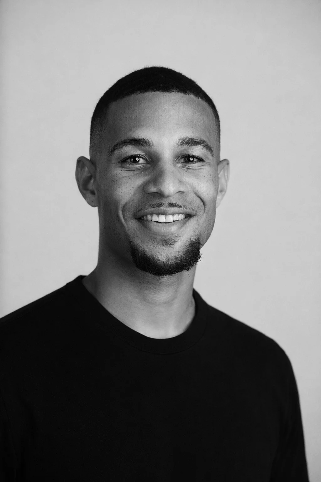 A man in a black quilted jacket and black hat speaking into a microphone indoors, with a window behind him showing a brick building and trees.