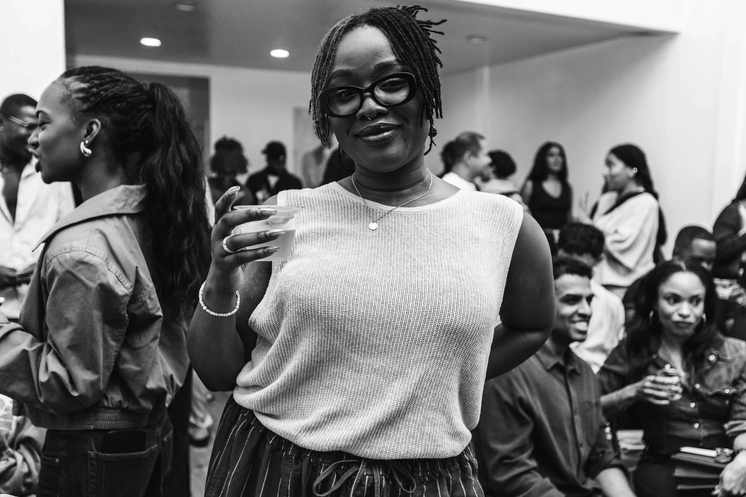 A woman with glasses and short, textured hair holds a drink at a social gathering, surrounded by other people engaging in conversation and enjoying the event.