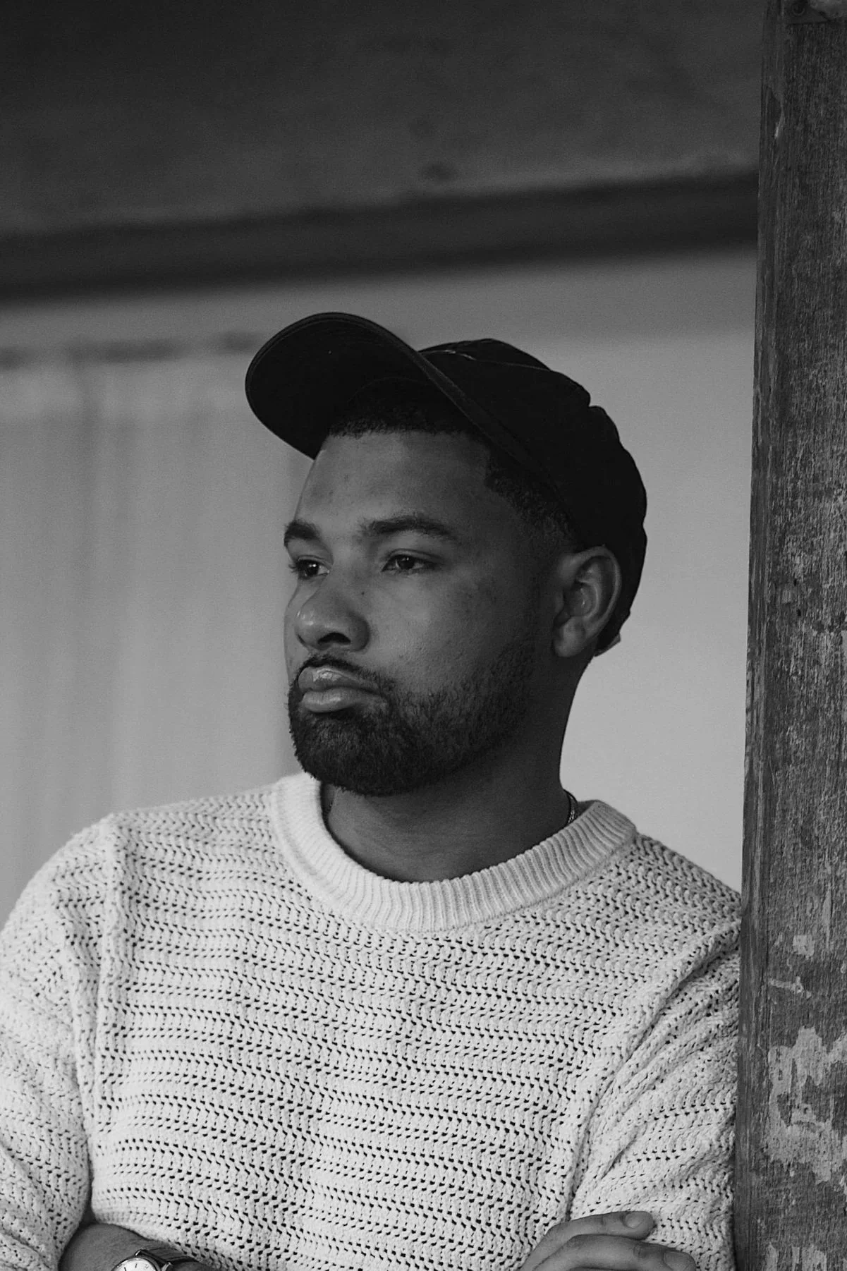A man with a beard wearing a beige knitted sweater and a dark cap, standing near a wooden post, looking thoughtfully into the distance.