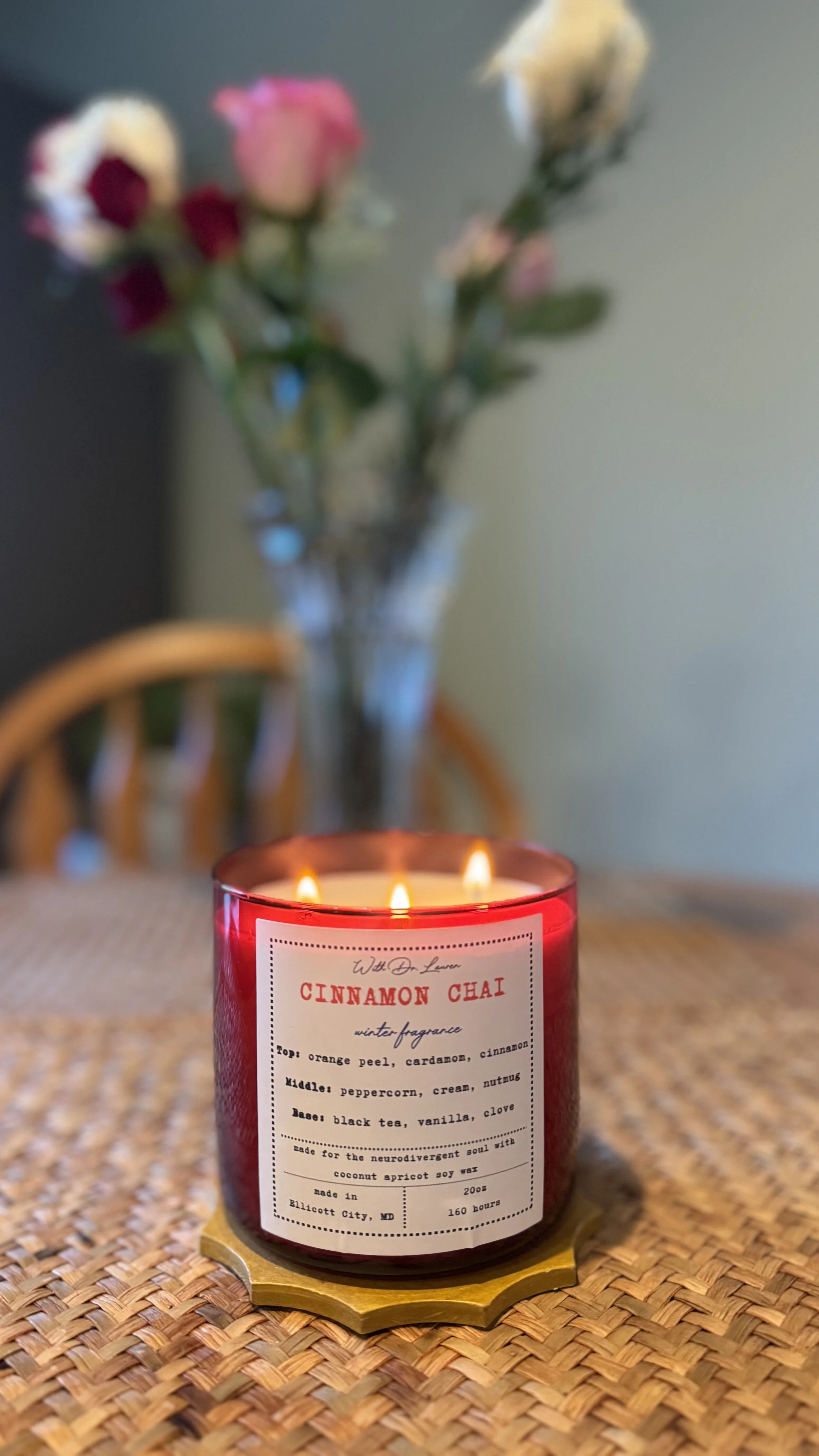 A lit cinnamon chai scented candle on a woven placemat with a vase of pink and white flowers in the background.