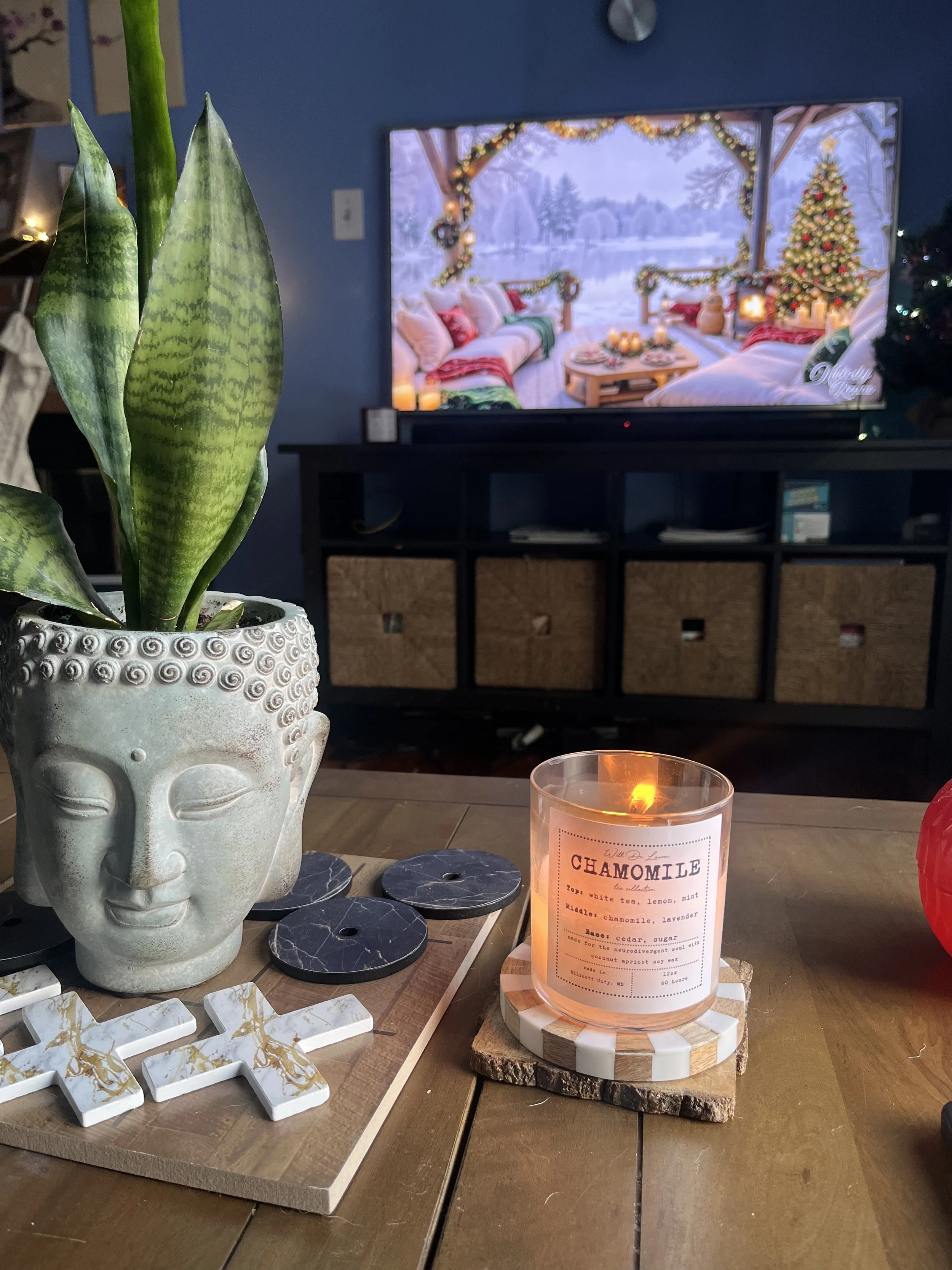 A decorative plant in a Buddha face-shaped pot, a lit candle labeled 'Chamomile', and cross-shaped decor on a table, with a television screen in the background displaying a cozy holiday scene with Christmas trees, pillows, and outdoor winter landscape.