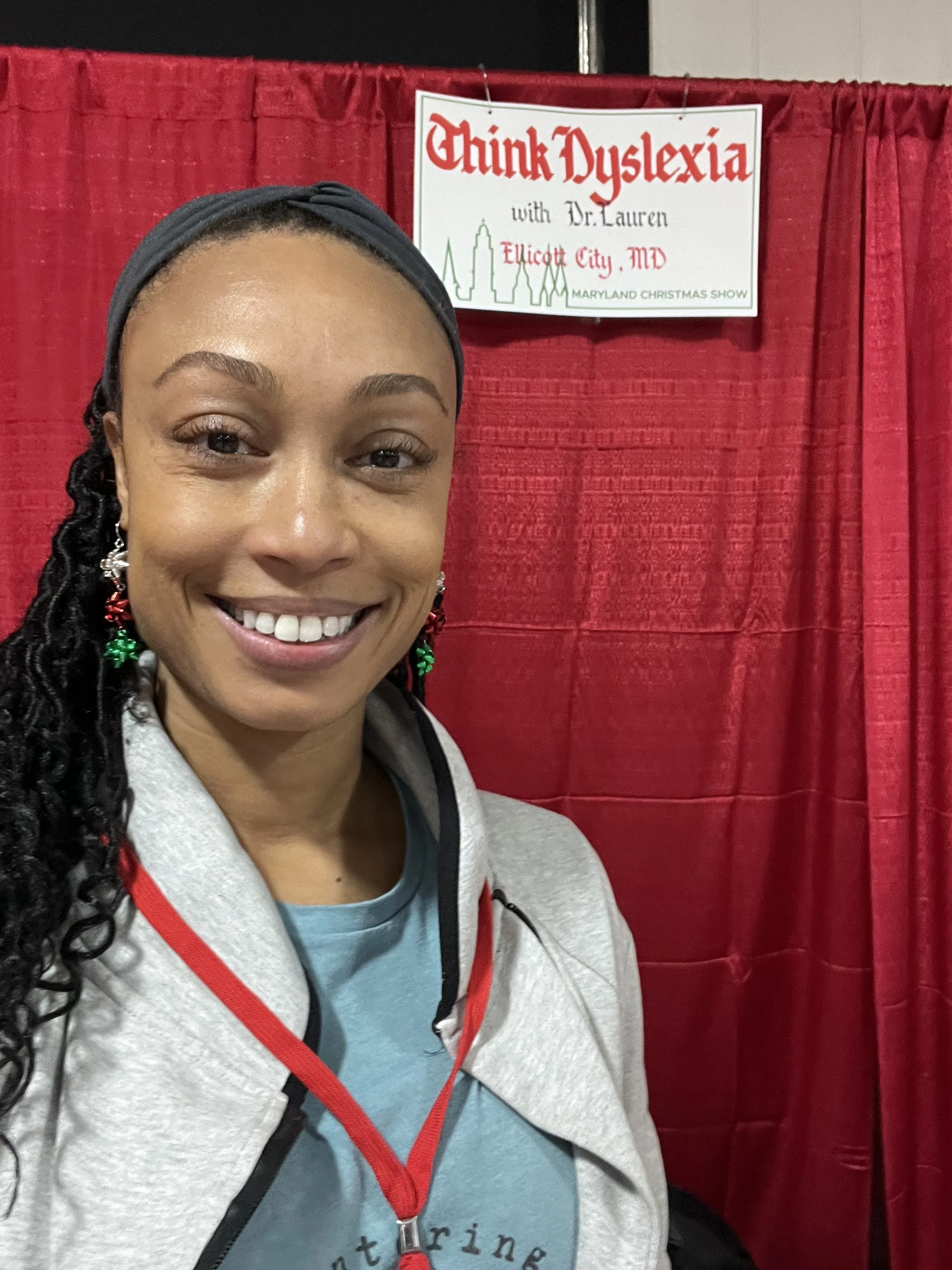 A woman smiling at a booth with a red curtain backdrop. A sign reads "Think Dyslexia with Dr. Lauren, Ellicott City, MD, Maryland Christmas Show."
