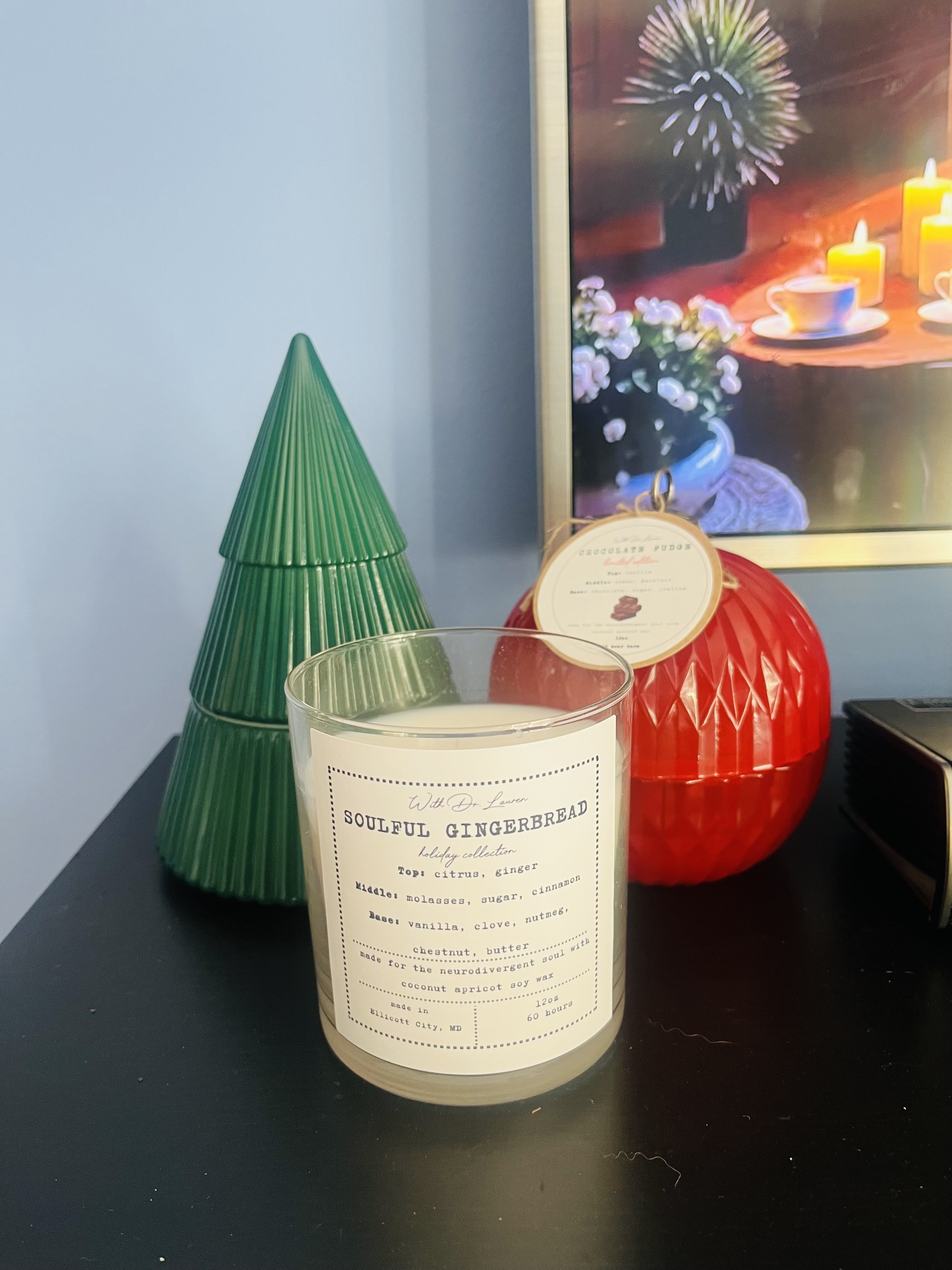 A holiday candle labeled 'Soulful Gingerbread' alongside decorative objects including a green Christmas tree, a red ornament, and a picture of candles and flowers in the background.