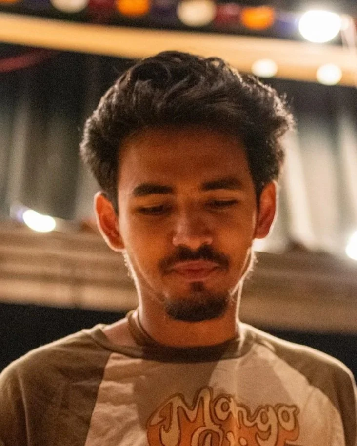 A young man with dark, wavy hair and facial hair, looking down, indoors with blurred background lights.