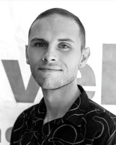Black and white photo of a man with short hair and a slight smile, wearing a patterned shirt, standing in front of a background with the letters 've' visible.