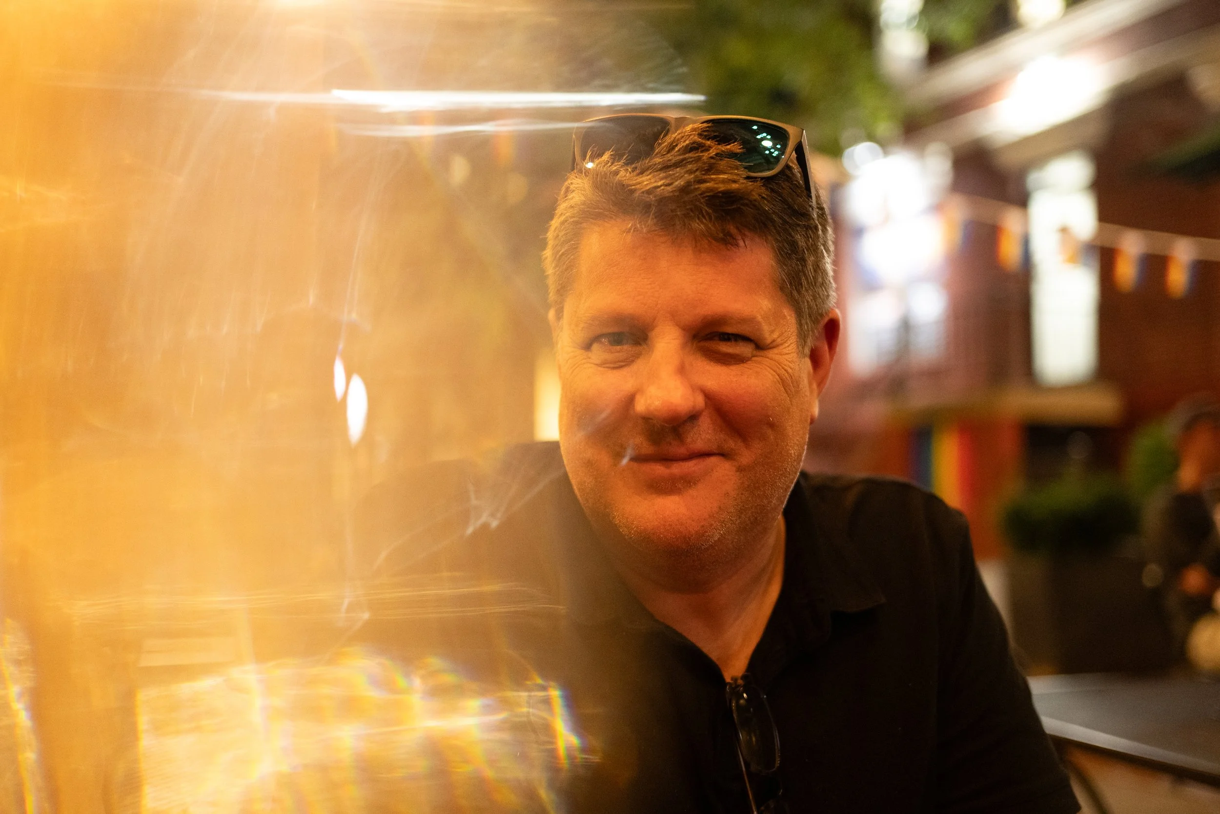 A middle-aged man with short gray hair and sunglasses on his head, smiling in a warmly lit, colorful indoor setting with shelves and decorations in the background.