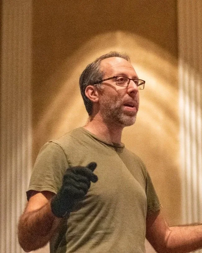 A man with glasses, a beard, and gray hair wearing a green t-shirt and black gloves, speaking or gesturing indoors with a decorative column and warm lighting in the background.