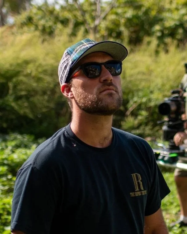 A man with a beard, wearing dark sunglasses, a plaid cap, and a black t-shirt, standing outdoors with a blurred green natural background and a person operating a camera in the distance.