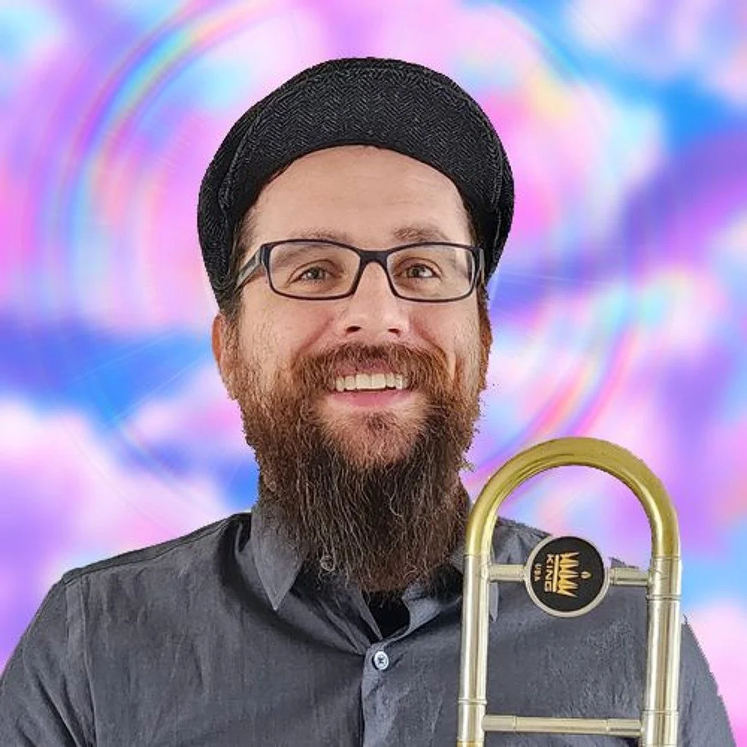 A man with glasses and a beard smiling, wearing a black hat and gray shirt, holding a trombone in front of a colorful, rainbow-like background.
