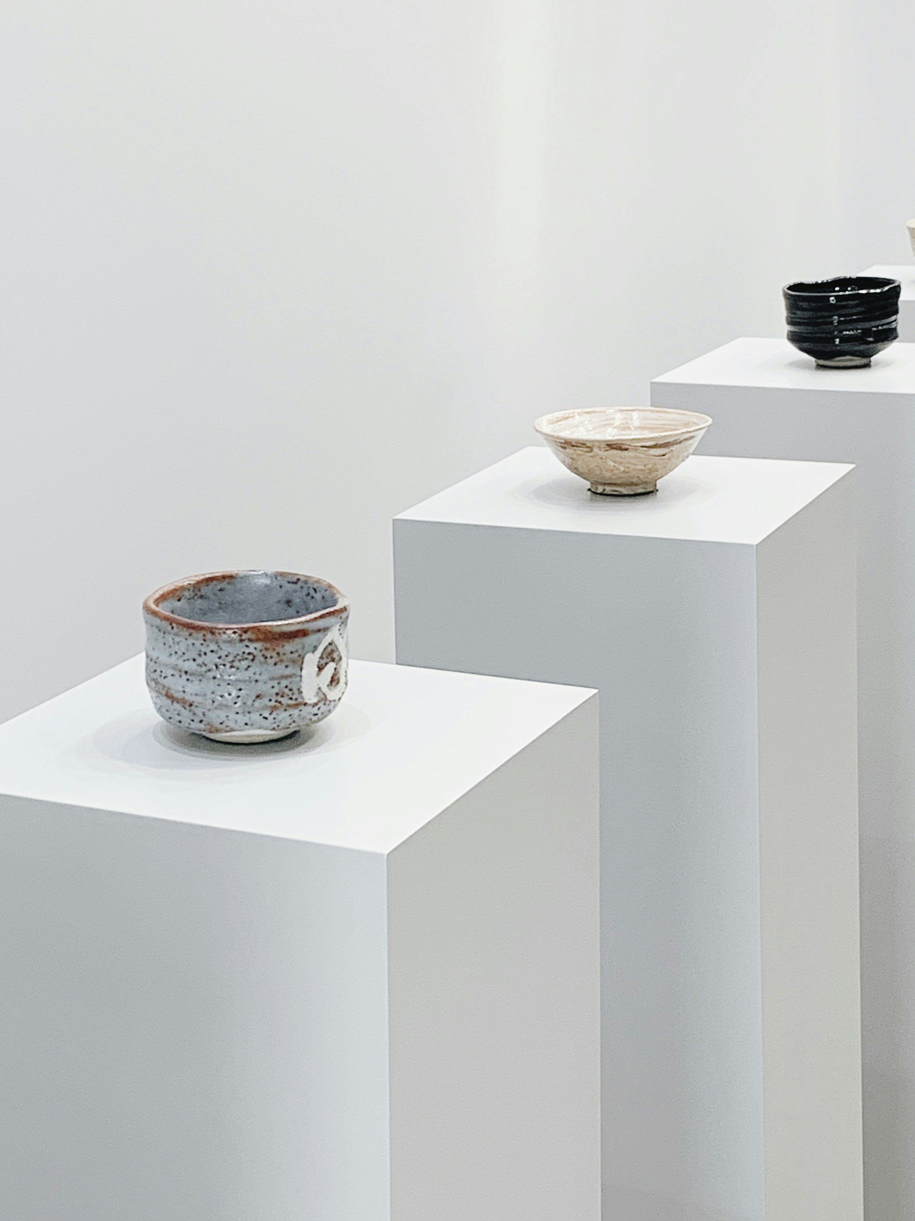 Three ceramic bowls displayed on white pedestals in an art gallery with a plain white wall background.