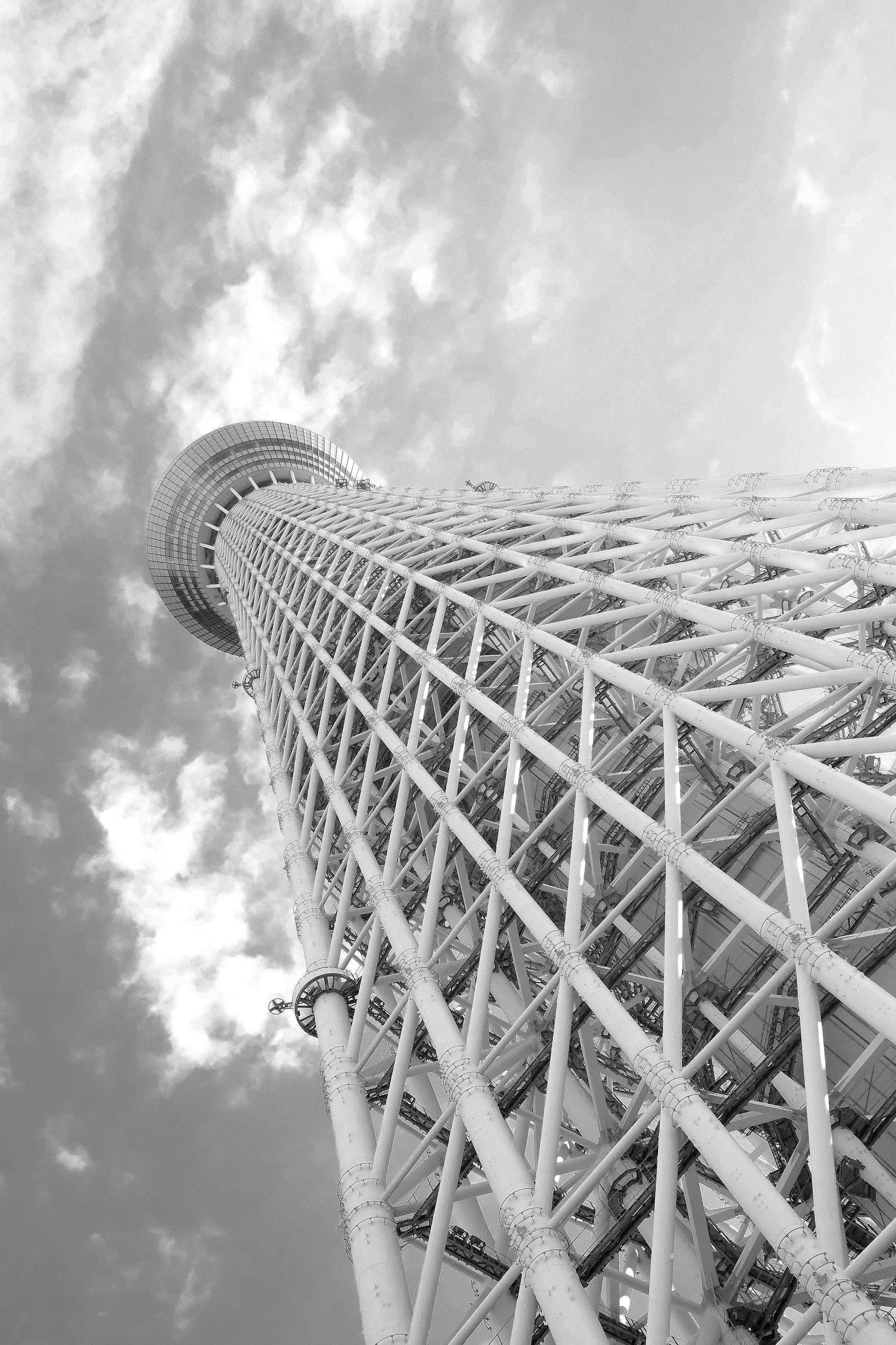 Low-angle black and white photo of a tall, cylindrical tower with a lattice structure and observation decks, set against a cloudy sky.