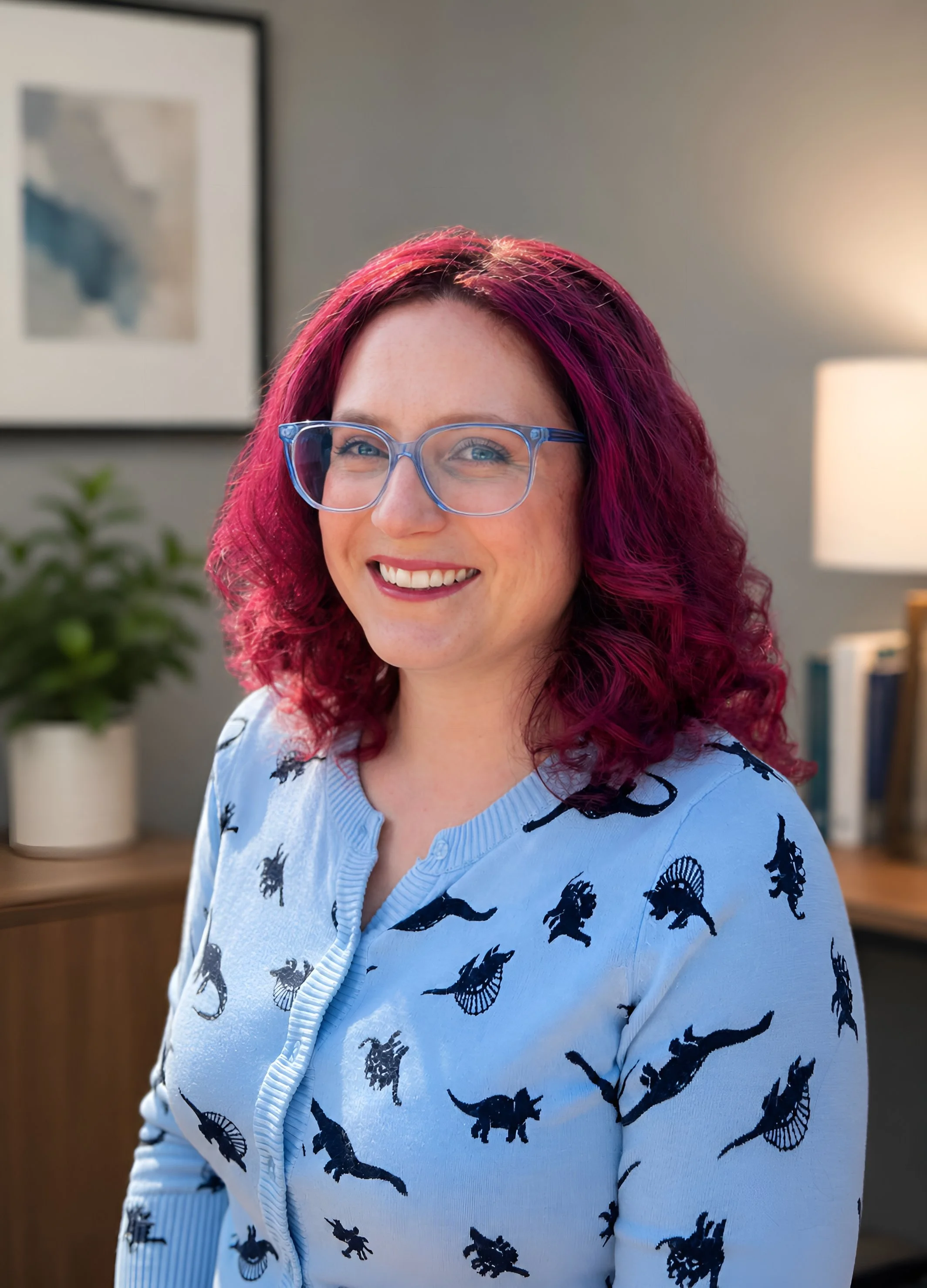 A woman with curly pink hair, glasses, and a light blue sweater with black insect patterns outdoors in a park.