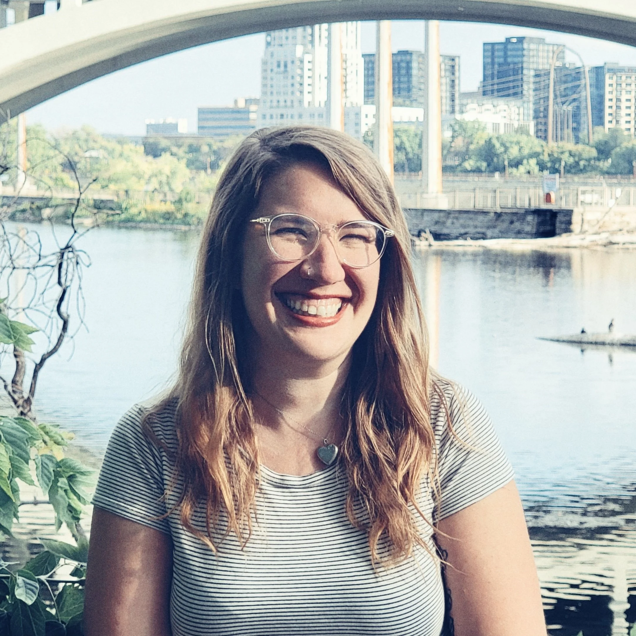 A woman with long, wavy hair and glasses smiling outdoors near a river with trees and city buildings in the background.