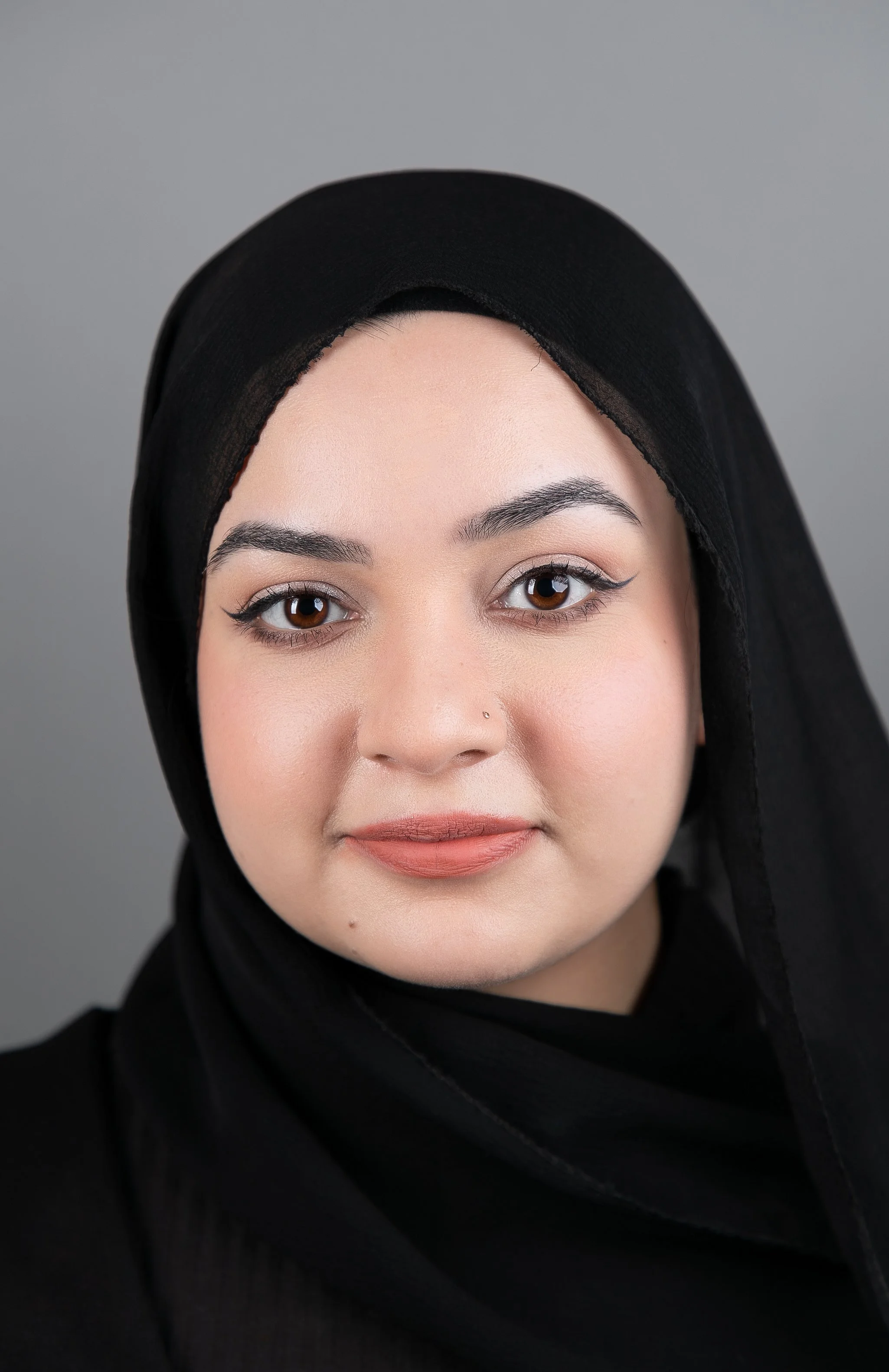 A close-up portrait of a woman with long brown hair and green eyes, wearing a black top, against a neutral background.