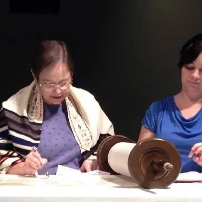 Two women sitting at a table with a large wooden spool on it, one older woman with glasses wearing a patterned shawl and a purple top, and a younger woman in a blue shirt.