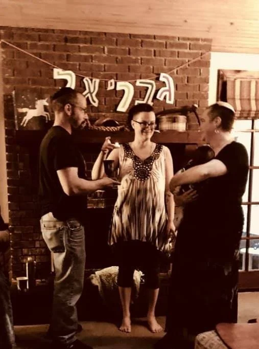 Three people standing and talking in a cozy living room with a brick fireplace and wooden mantel, decorated with a string of Jewish symbols and a silhouette of a dog, possibly celebrating a Jewish occasion.