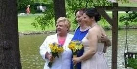 Three women standing outside near a pond, holding bouquets of flowers, enjoying a sunny day.