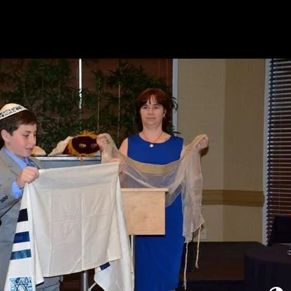 A woman stands behind a wooden platform with a young boy to her left, holding a tall white cloth. The woman wears a blue dress, while the boy wears a kippah and a white tallit with blue stripes. They are indoors, near a window with blinds, with plant