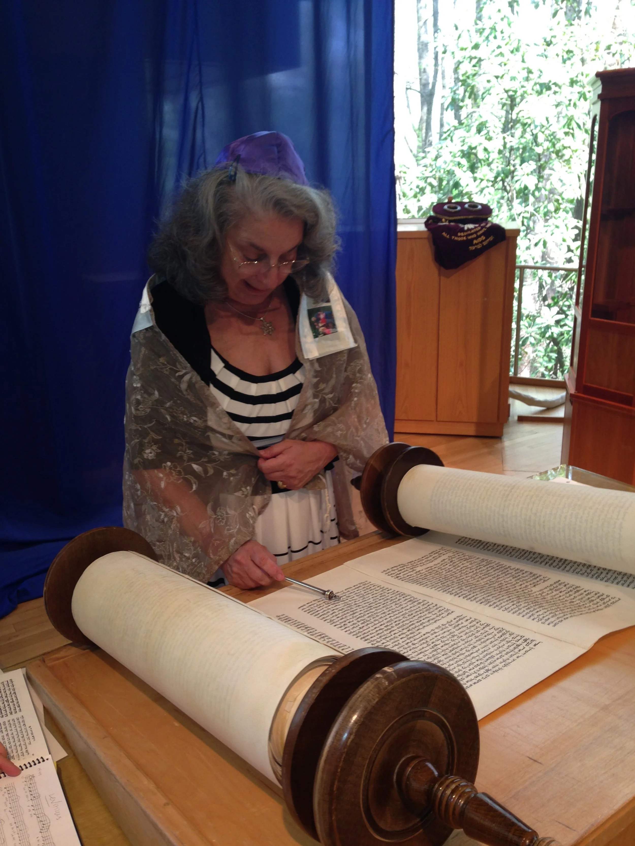 An elderly woman wearing a striped shirt and a lace shawl is looking at a Torah scroll with a pointer in her hand. She has gray hair and is wearing glasses, standing in a room with a window showing greenery outside.
