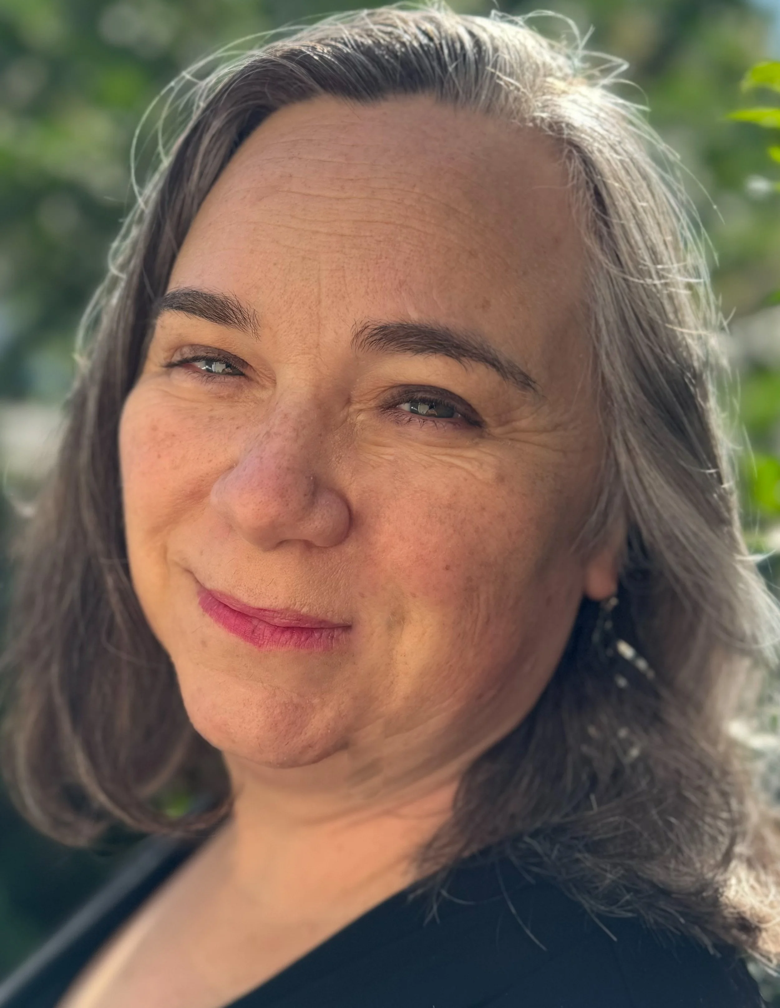 Close-up photo of a woman with light skin, brown hair, and faint freckles, smiling outdoors with a blurred green background.
