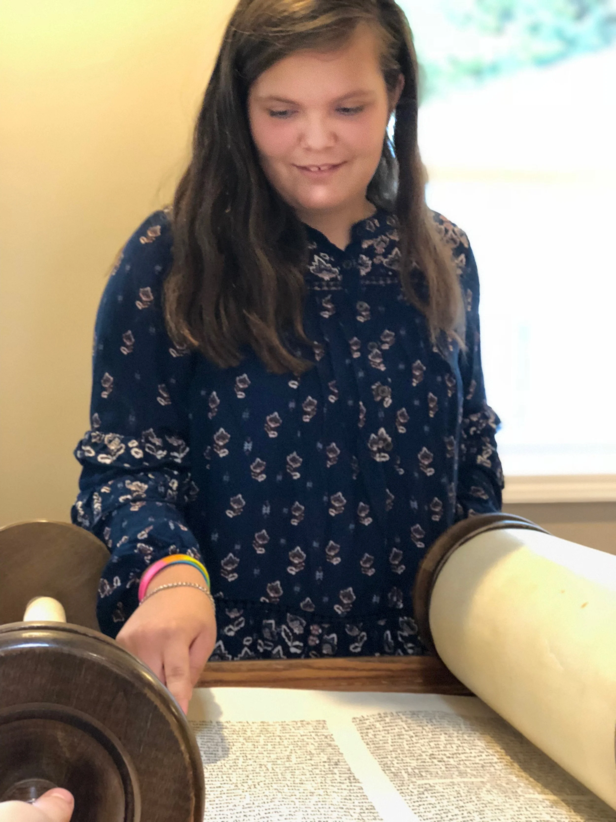 A young woman with long brown hair, wearing a navy blue patterned blouse, looks down at a large scroll and wooden device on a table in a room with a yellow wall and a window in the background.