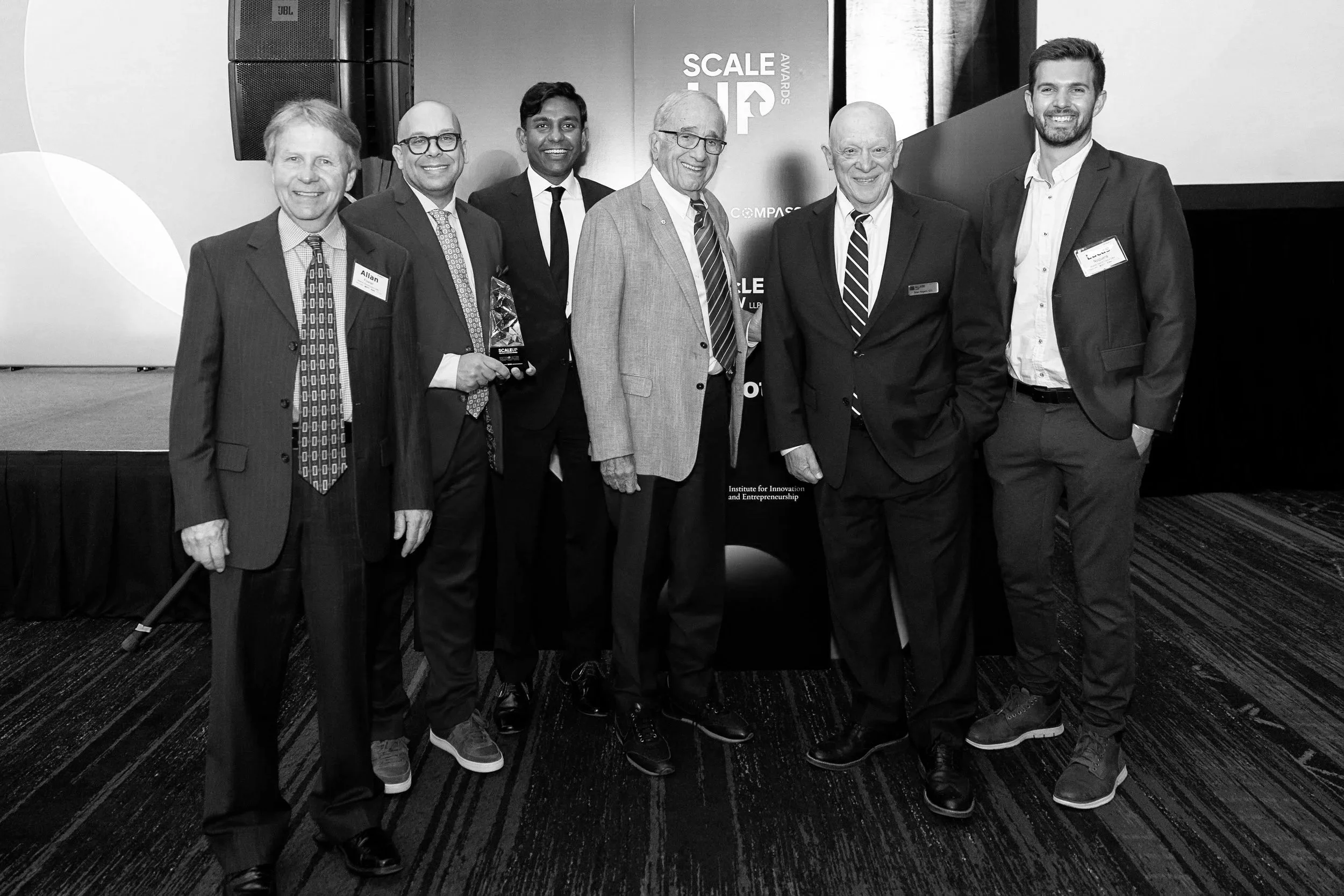 Black and white photo of seven men in business suits standing together at an event, smiling at the camera. One man holds a trophy. Behind them are banners with event branding.