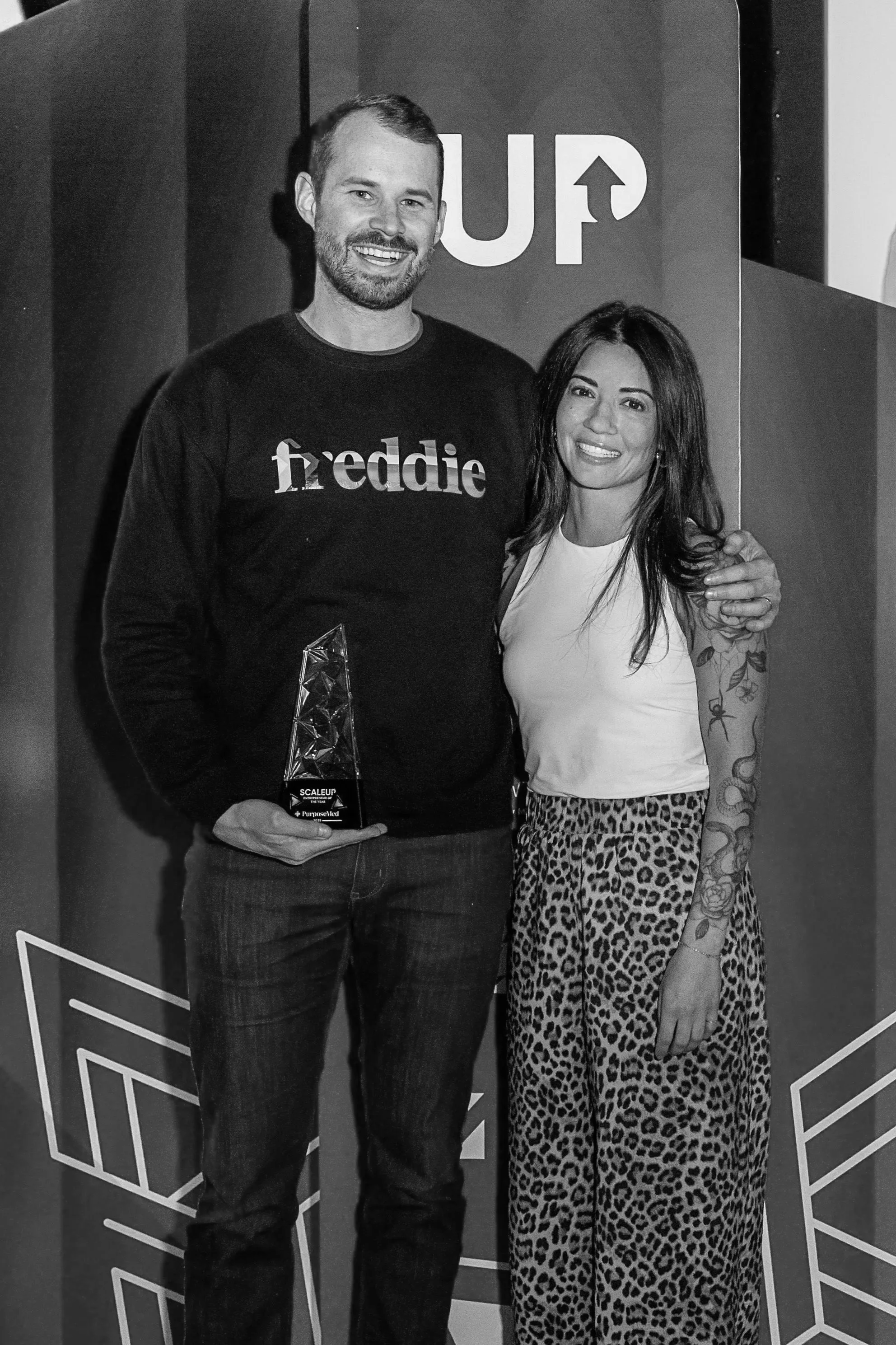 A man and woman standing together at an award ceremony, smiling for the camera. The man is holding a trophy in his left hand. The background features a sign with partial letters 'UP' and geometric designs.