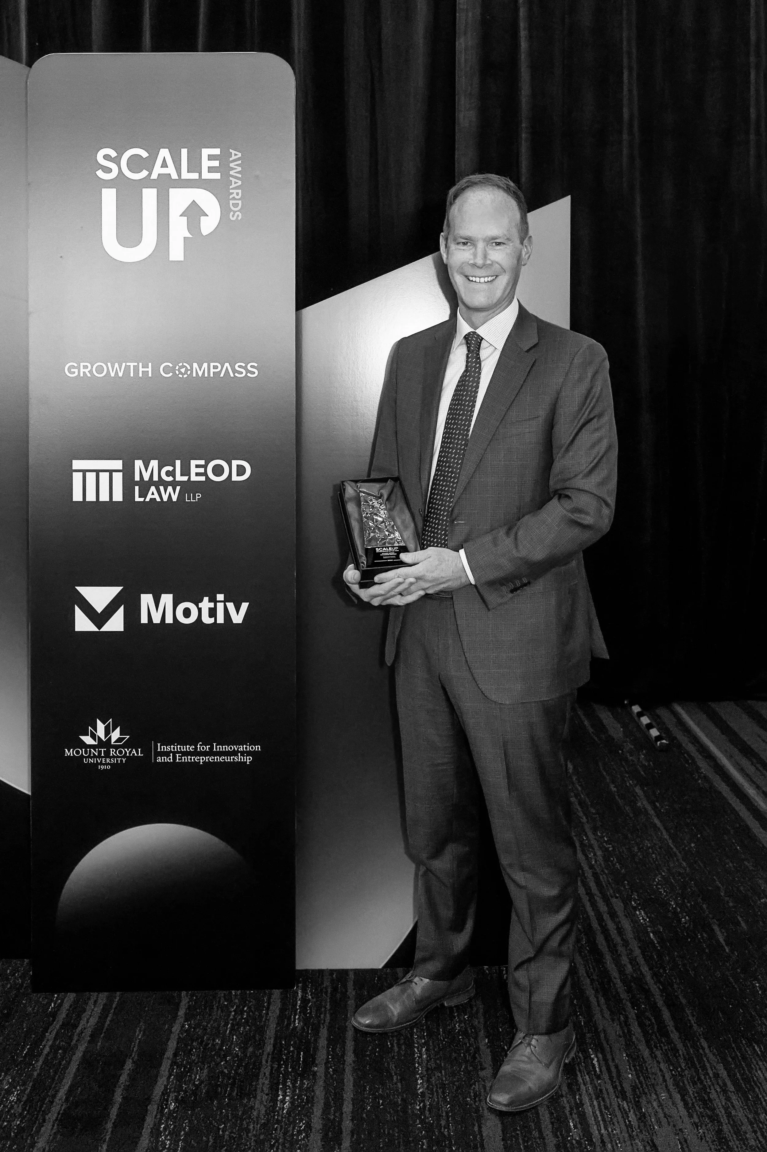 A man in a suit holding an award standing next to a vertical banner with logos of various organizations and event titles at a conference or award ceremony.