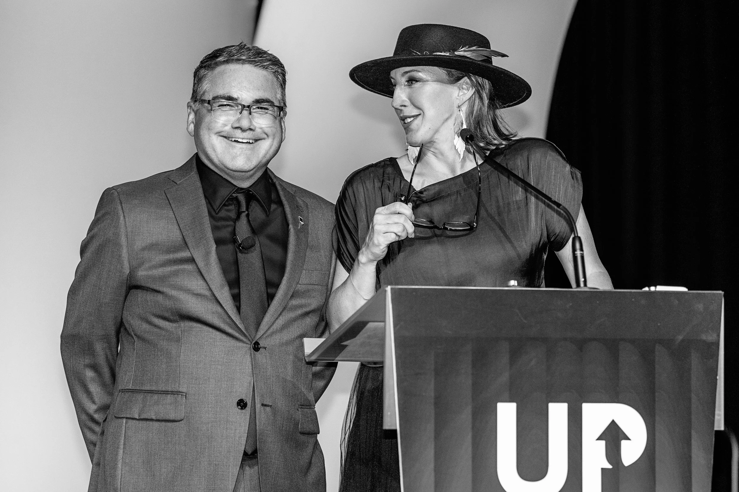 Two people standing at a podium, one man in a suit and a woman wearing a hat, speaking at an event.