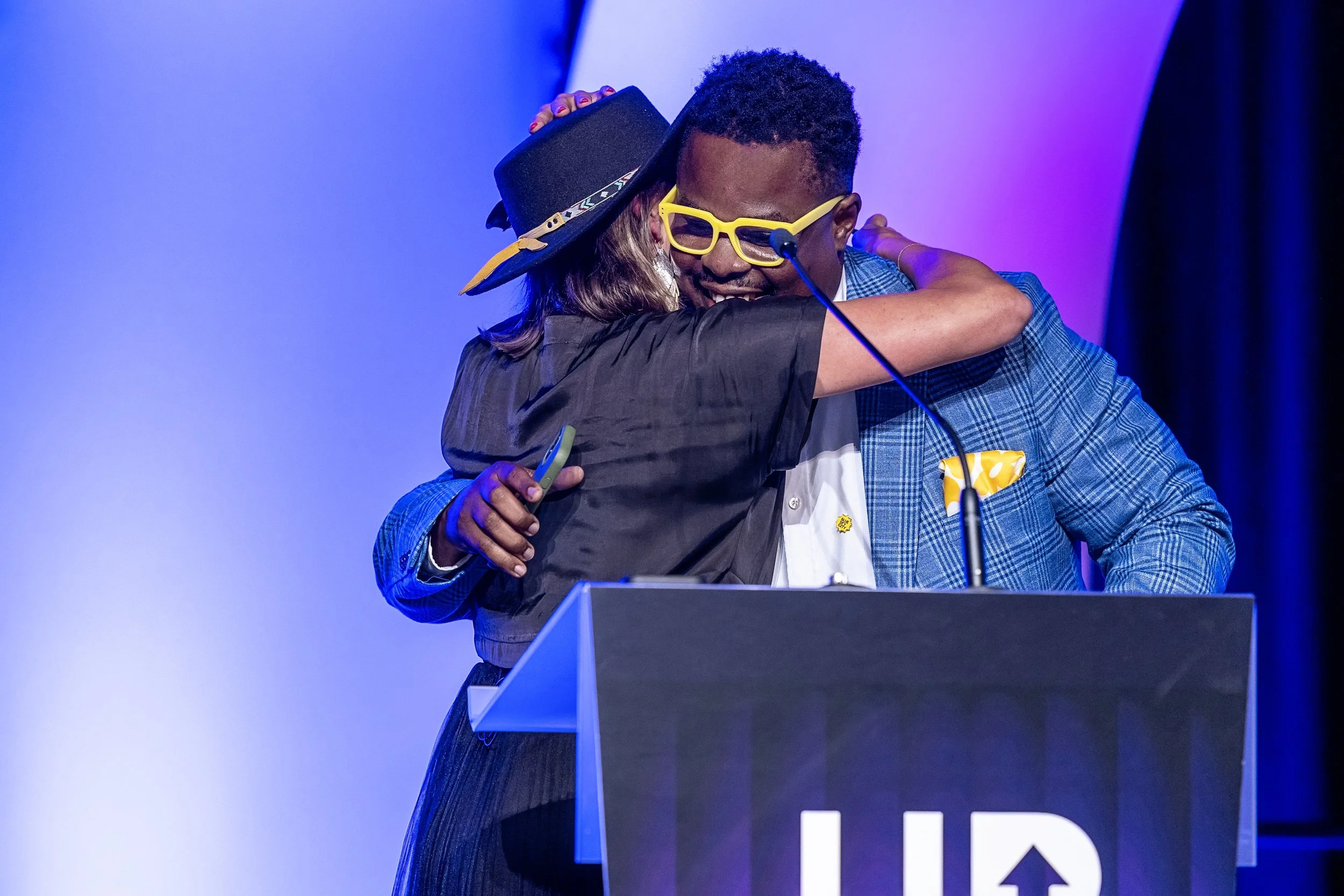 Two people hugging at a podium with a partially visible logo, one wearing yellow glasses and the other wearing a black hat, with colorful lighting in the background.