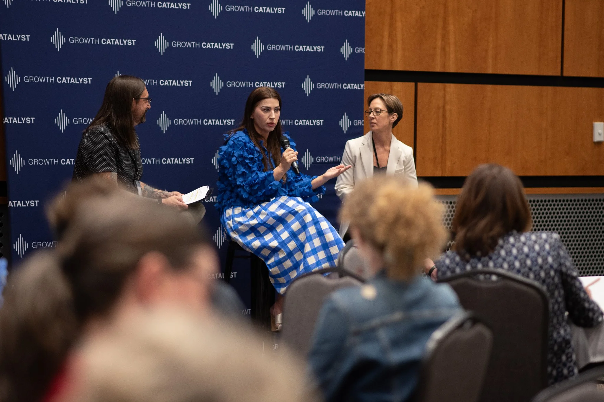 Three women seated on stage during a panel discussion at an event with a blue backdrop displaying 'GROWTH CATALYST' in white text and a logo. The woman in the center, holding a microphone, wears a blue textured top and a blue and white checkered skirt. The woman on the right wears a white blazer, and the woman on the left has long hair and glasses, holding a paper.